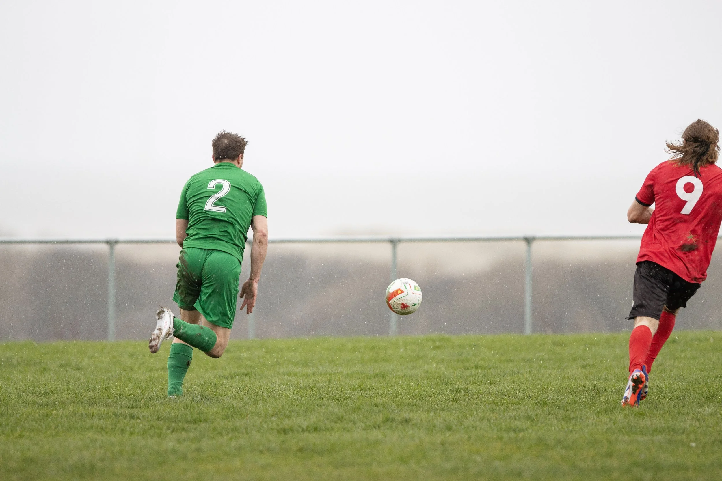 Two soccer players, one in a green jersey with number 2 and the other in a red jersey with number 9, chasing a soccer ball on a grassy field during a rainy day.