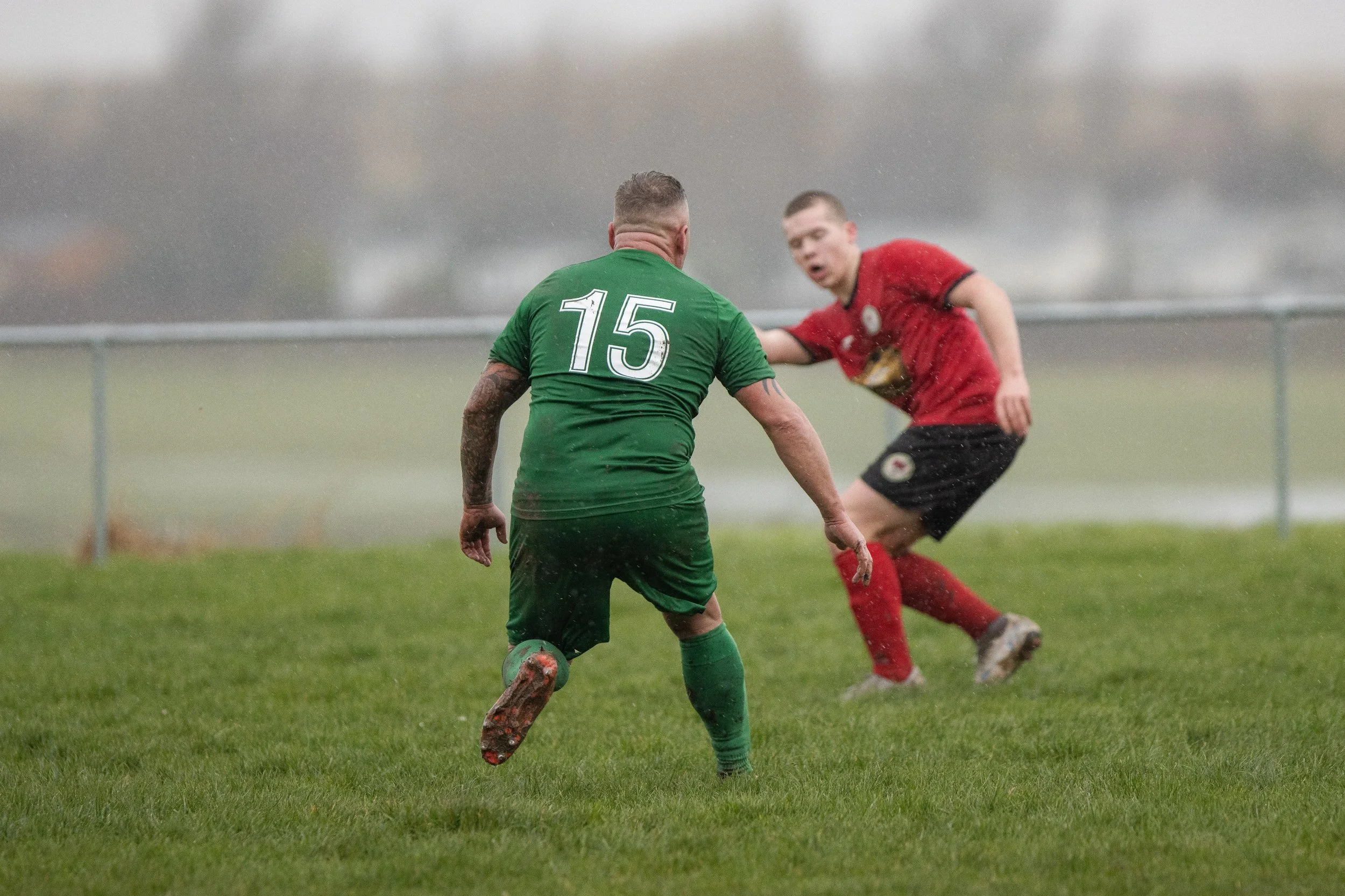 Two soccer players on a wet field during a game, one in a green jersey with number 15 and the other in a red jersey. The player in red appears to be in motion while the green jersey player is moving forward.