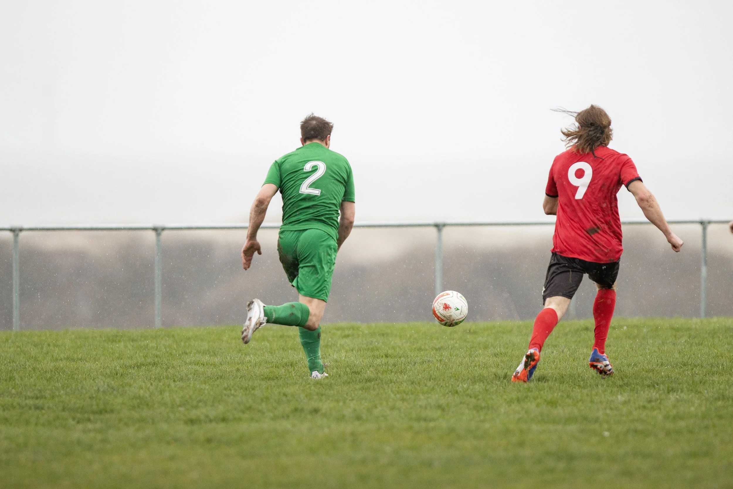 Two soccer players running after a ball on a grassy field, one in a green jersey with number 2 and the other in a red jersey with number 9.