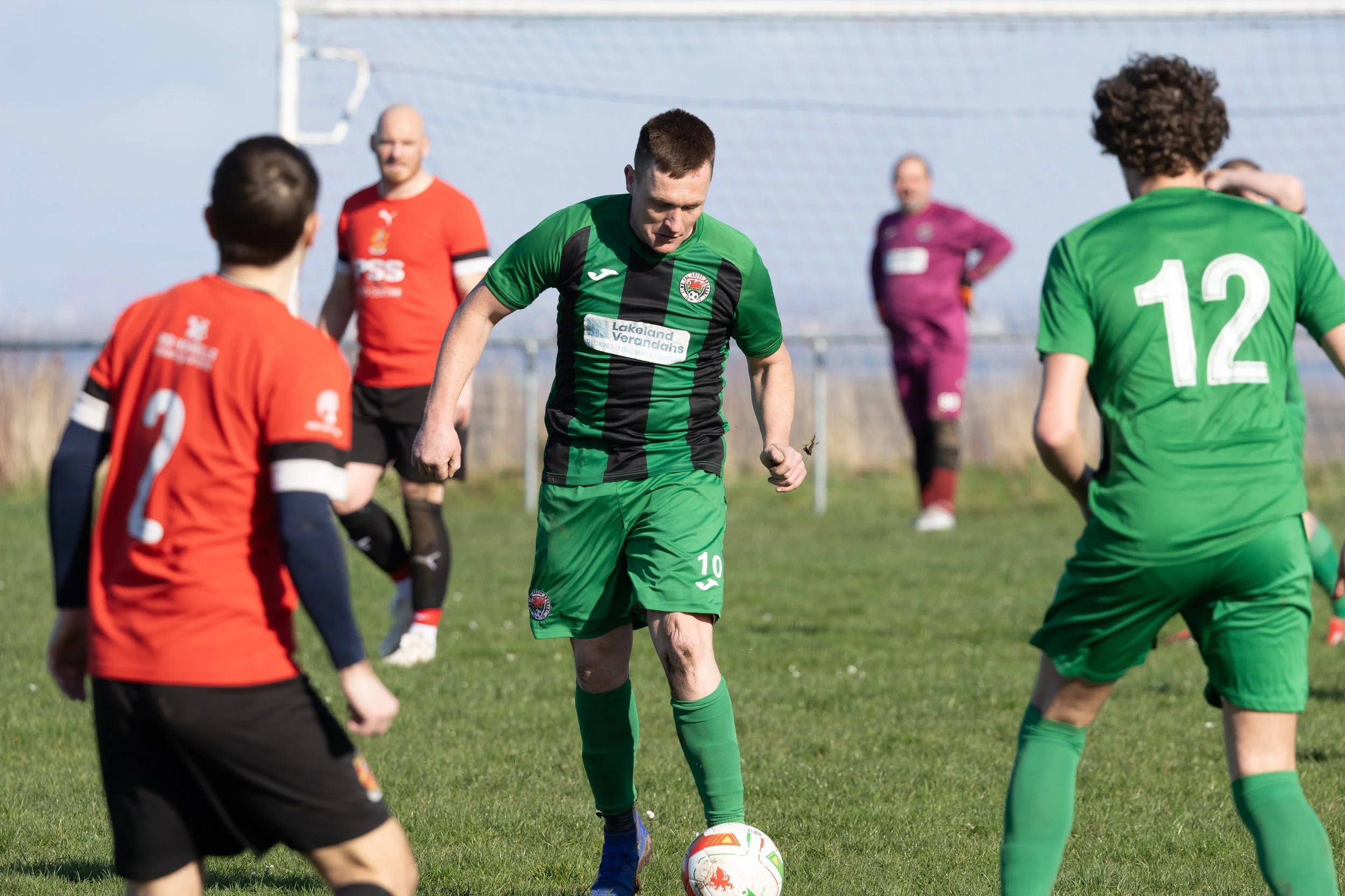 Soccer players on a field during a match, with one player in green and black about to kick the ball, and others in red and green jerseys nearby.