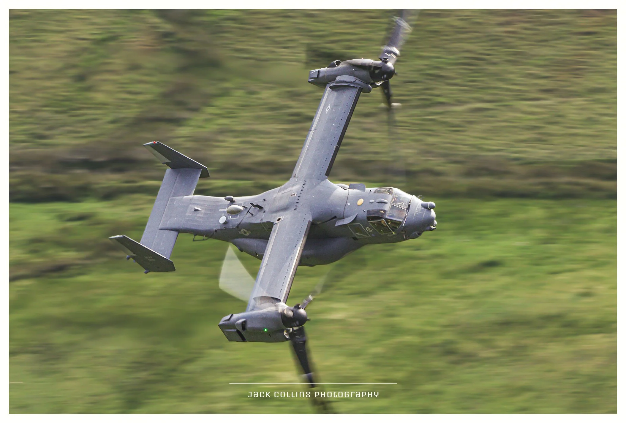 A military aircraft flying at an angle against a blurred green landscape background.