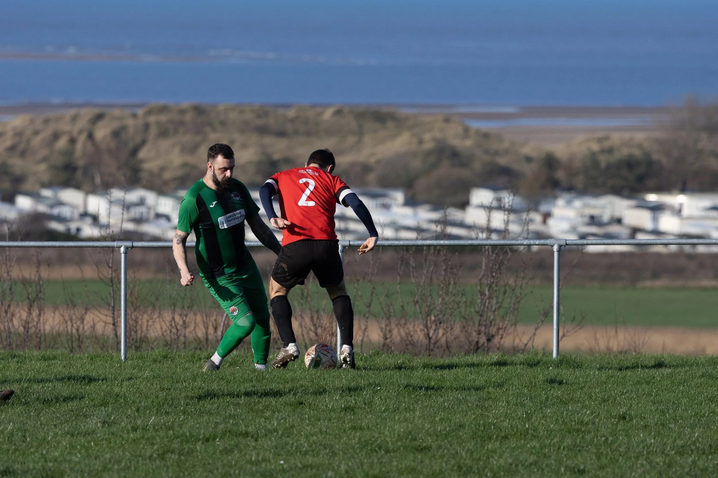 Two soccer players compete for the ball on a grassy field, with a rural landscape and water in the background.