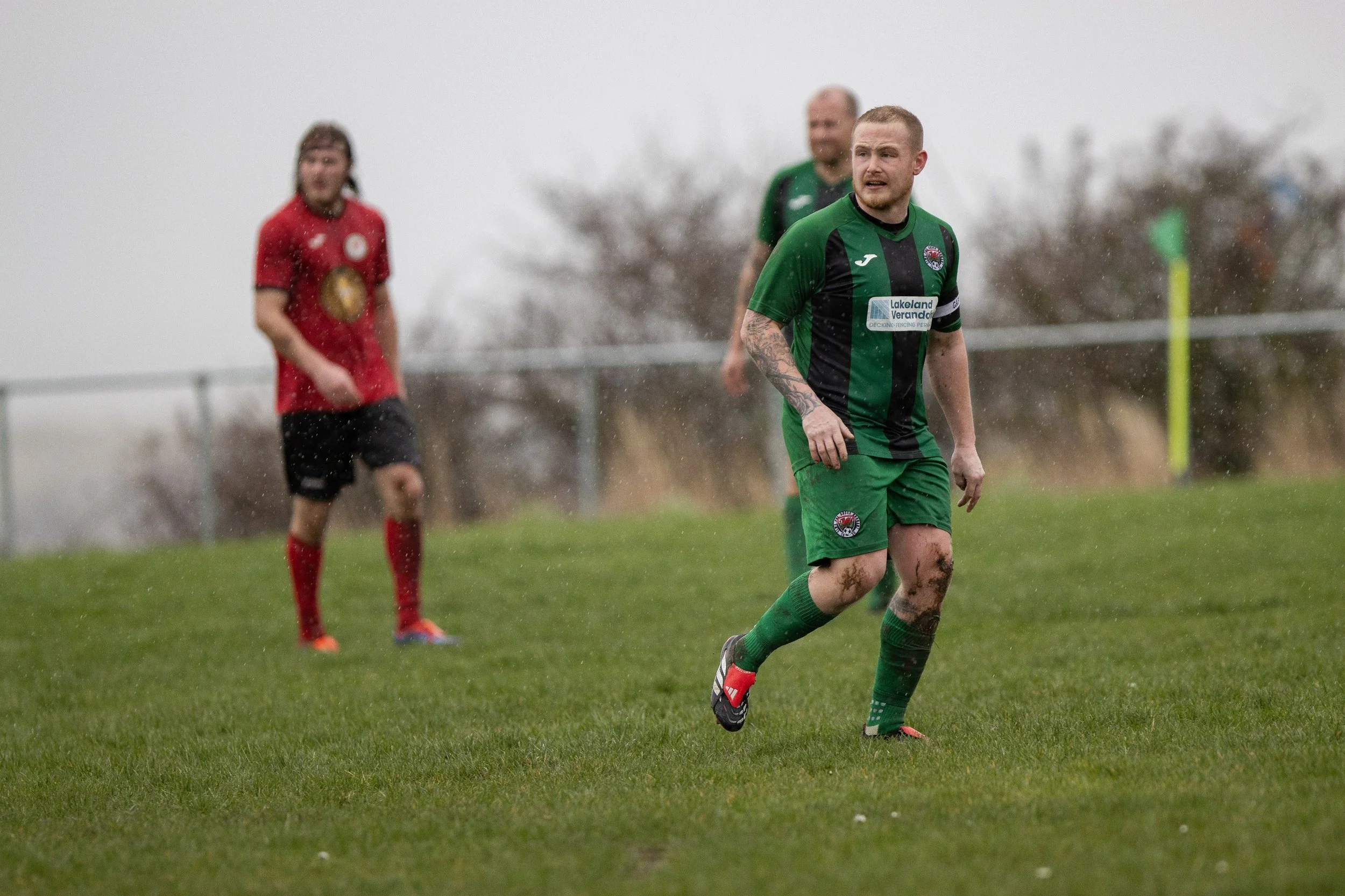 Soccer match on a rainy day with players wearing green and red jerseys on a muddy field.