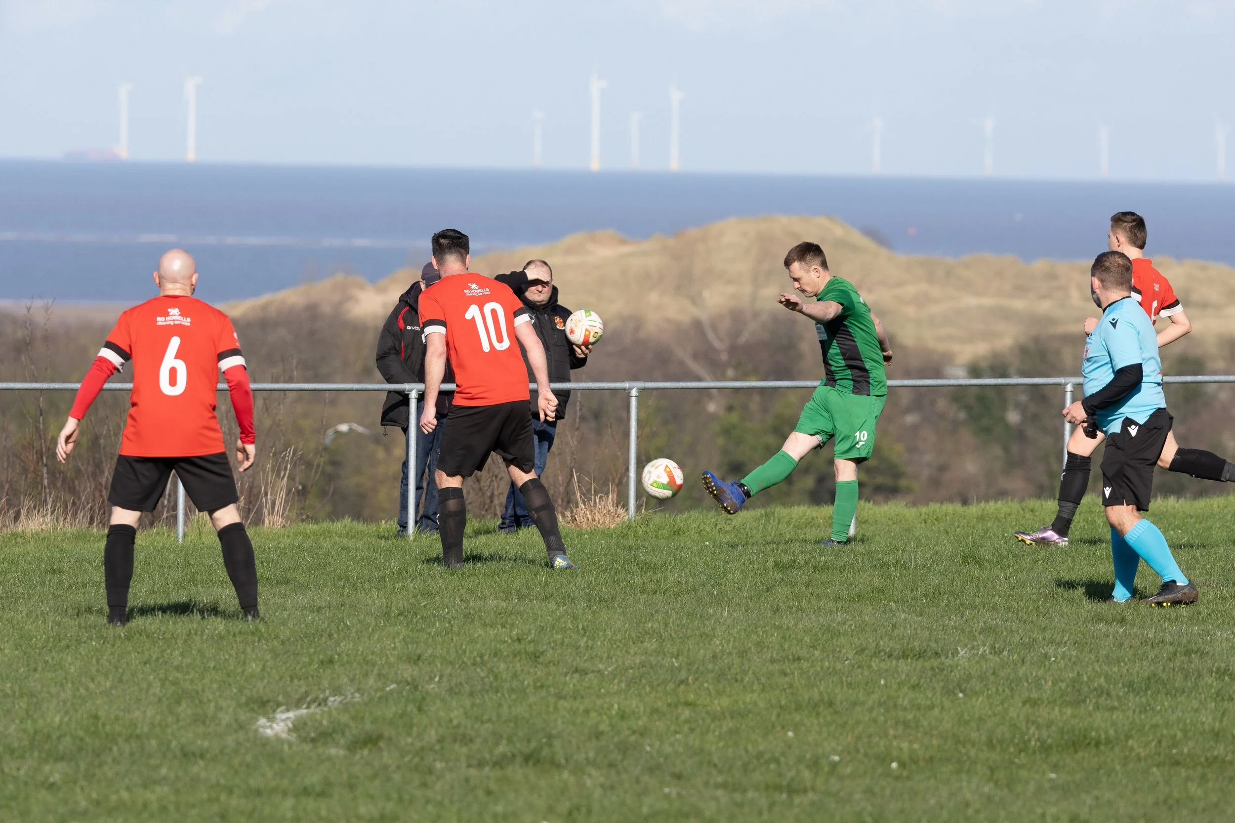 Soccer players on a field during a match, with one player in a green uniform kicking the ball while others in red and black uniforms observe, and a referee in a light blue shirt. In the background, there are hills and wind turbines.