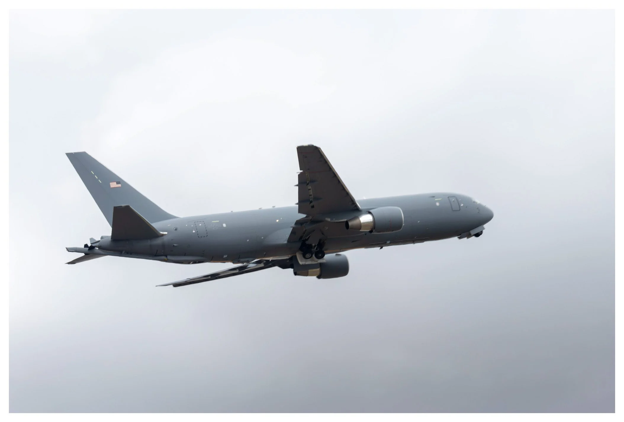 A military aircraft flying through a cloudy sky, painted in dark gray with a U.S. flag on the tail.