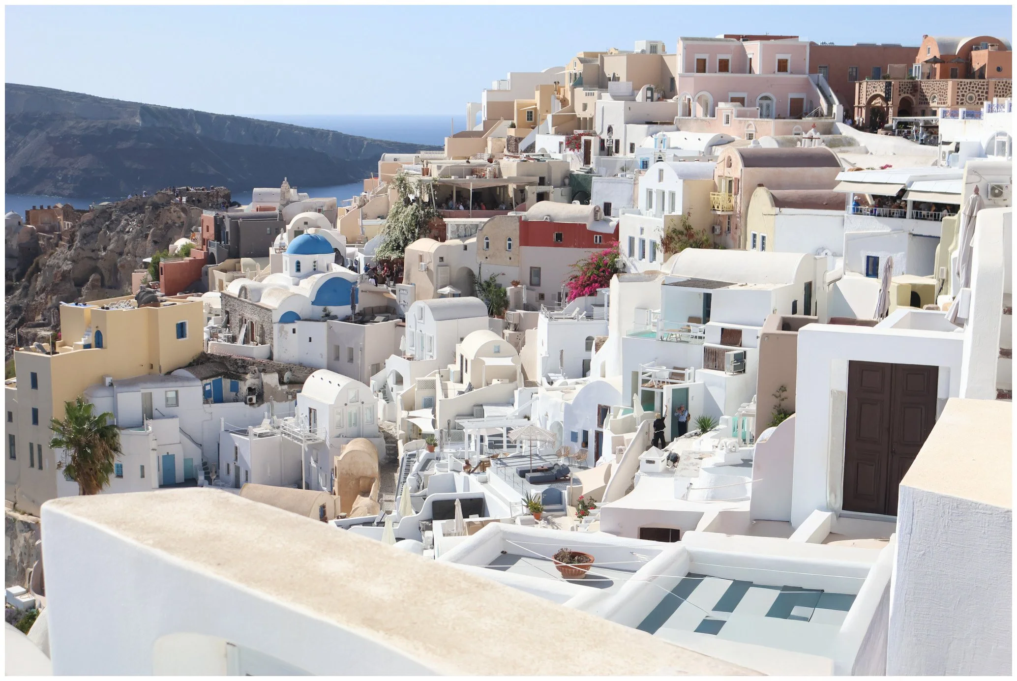 The image features a scenic view of the white-washed buildings of Santorini, Greece, with the Aegean Sea and cliffs in the background. The architecture includes domed roofs and narrow pathways.