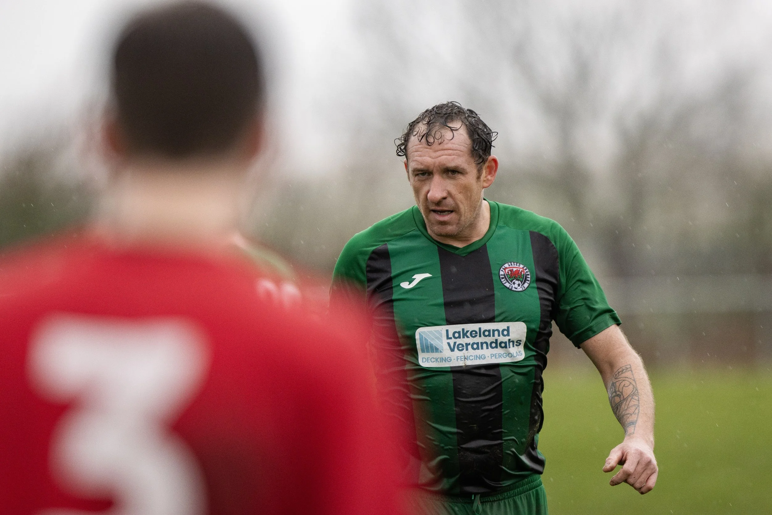 A soccer player with wet hair and a tattoo on his left arm, wearing a green and black uniform, is looking at another player in a red jersey during a game on a rainy day.