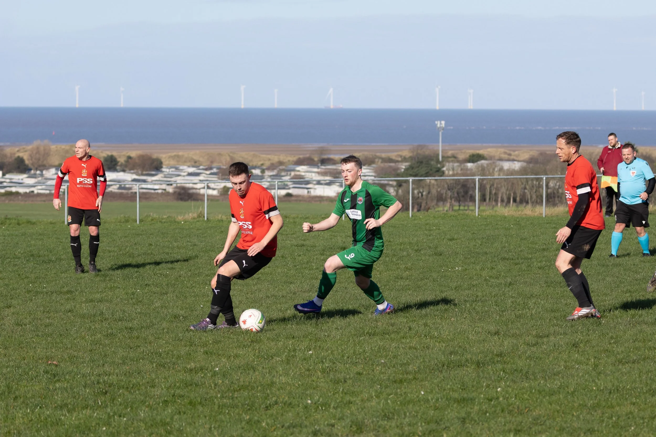 Soccer game on a grassy field with players in red and green jerseys, and a referee in turquoise in the background, near a fence with a view of the ocean and wind turbines in the distance.