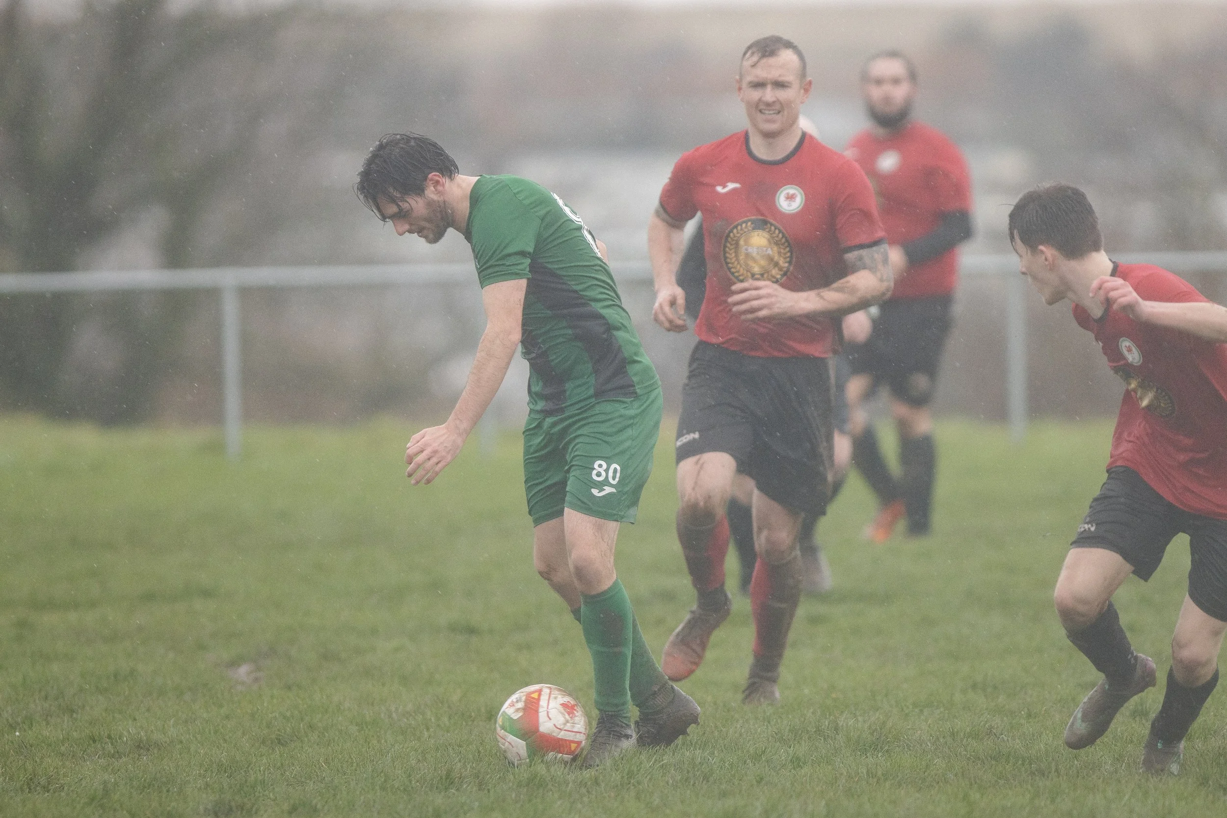 Soccer players playing on a rainy field, with one player in a green uniform controlling the ball while others in red jerseys run behind him.