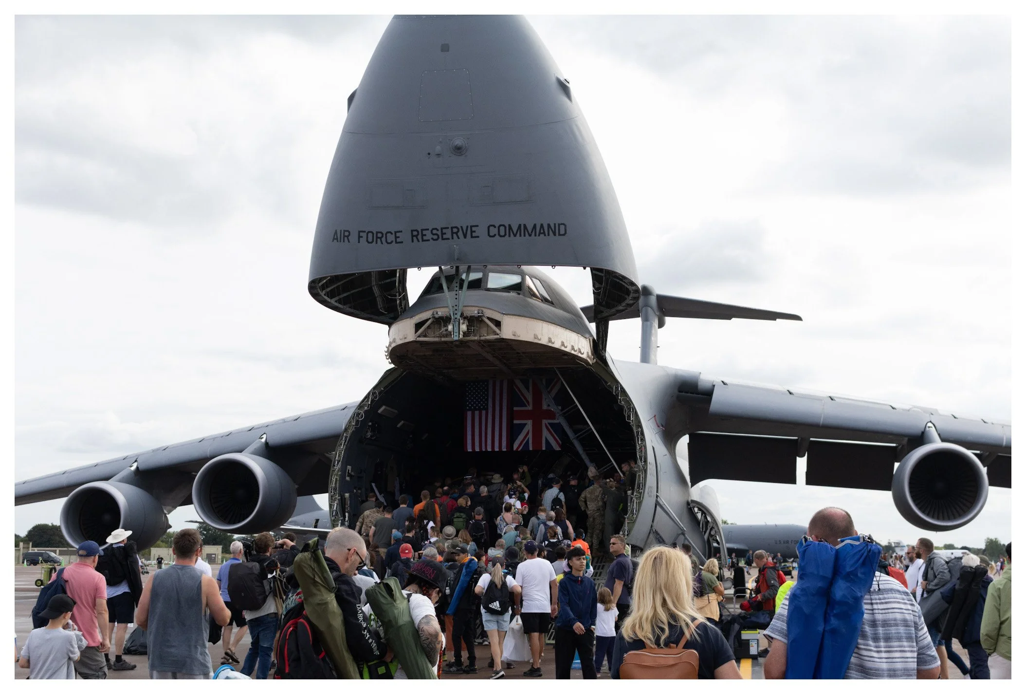People boarding a large military cargo plane with the rear doors open, inside some flags are visible, including the American and British flags, and the top of the plane has the words 'Air Force Reserve Command'.