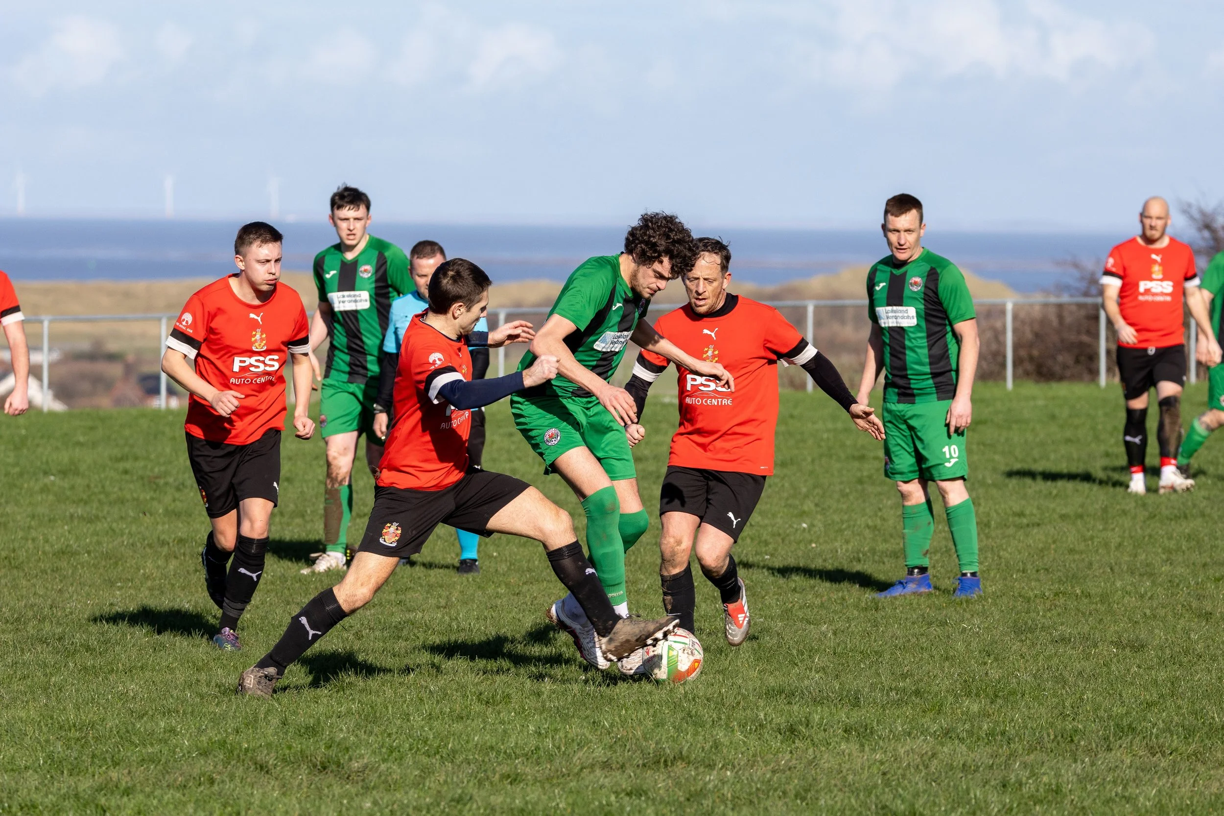 Soccer match with players in red and green uniforms competing for the ball on a grassy field with a fence and scenic landscape in the background.