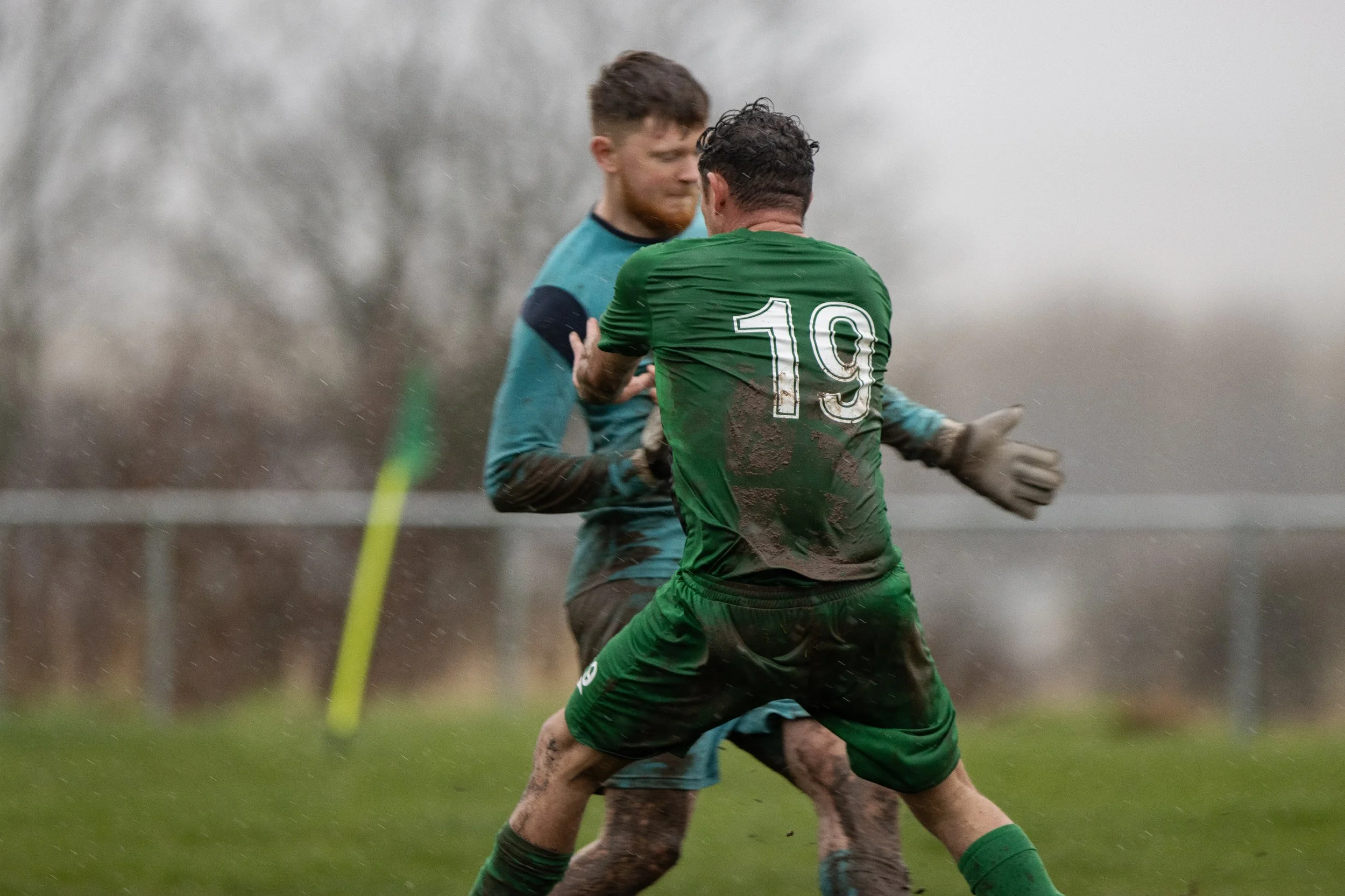 Two soccer players in muddy uniforms, one with the number 19, are engaged in a game on a rainy day.