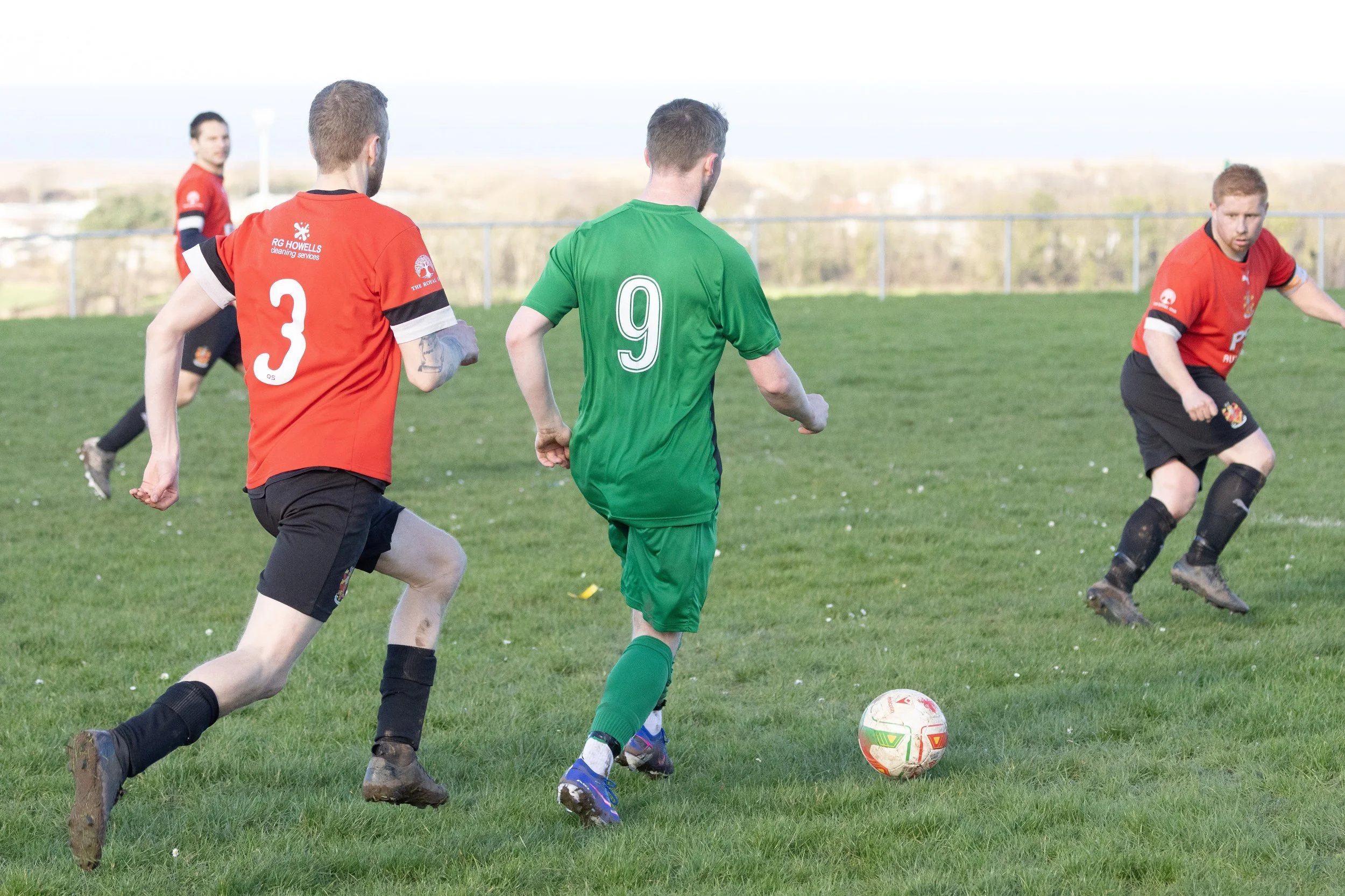 Soccer players competing on a grassy field, with one in green and others in red jerseys, chasing the ball.