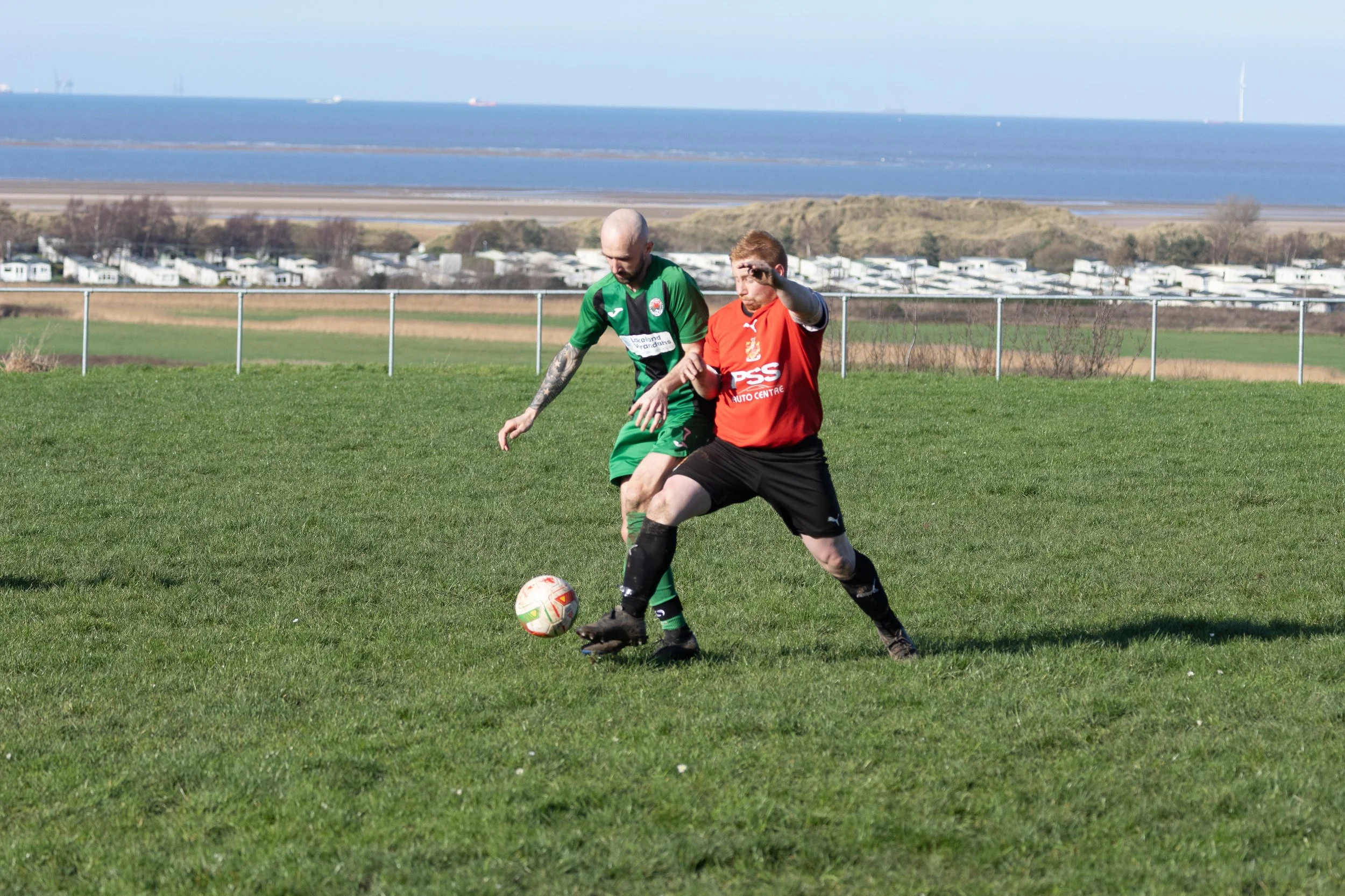 Two soccer players competing for the ball on a grassy field with a fence, ocean, and boats in the background.