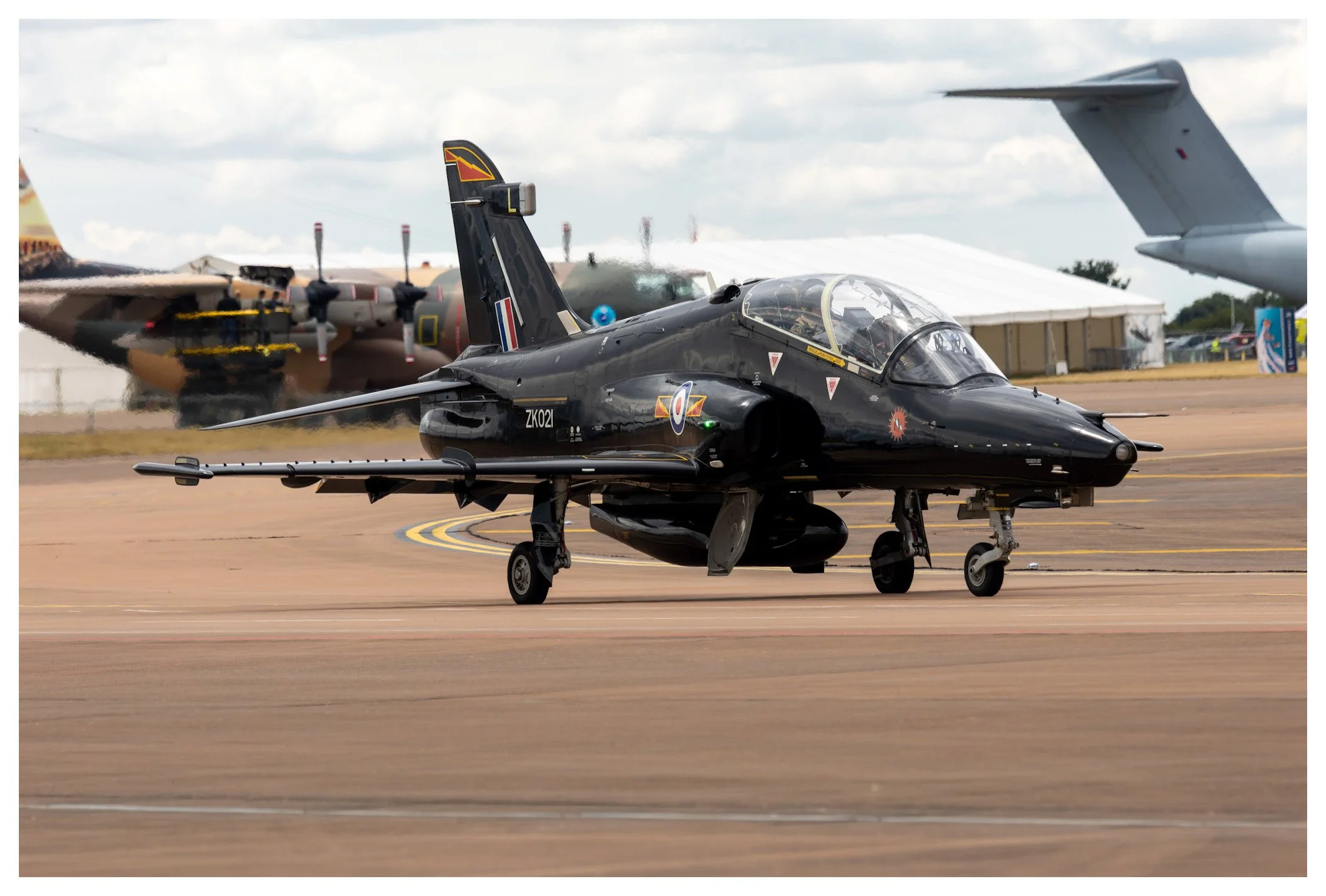 A black military jet aircraft on the runway with other planes and a hangar in the background at an airshow.