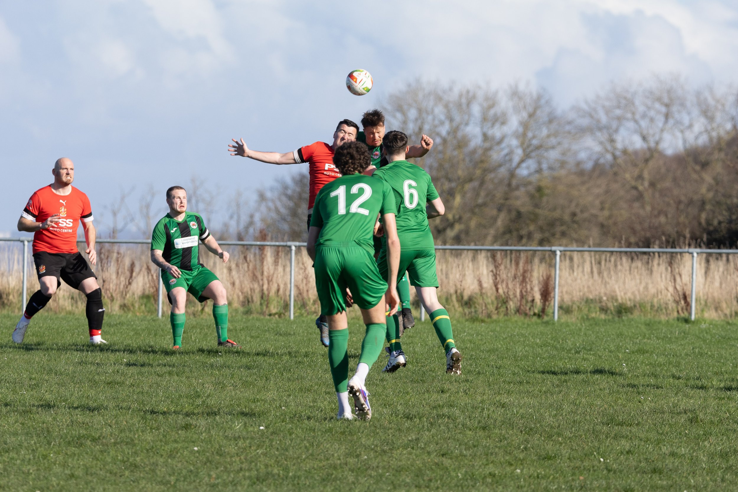 Soccer players in green and red uniforms jump and run on a grassy field during a match, with a ball in mid-air.