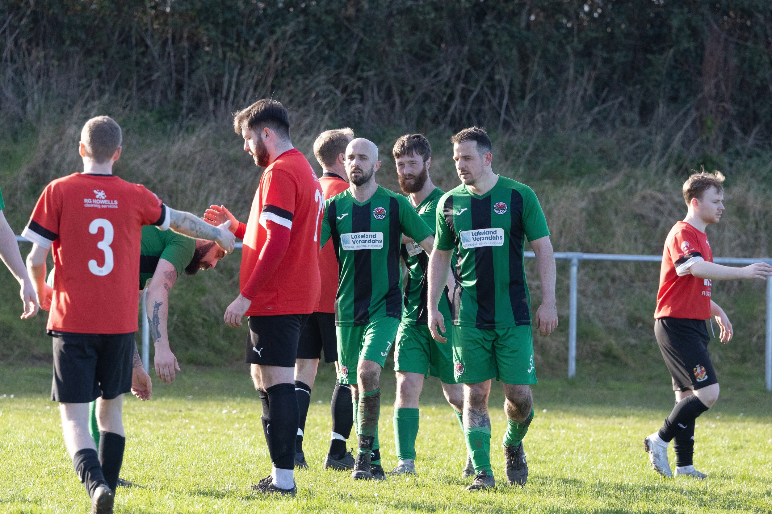 Soccer players in green and red jerseys on a grassy field exchanging handshakes after a match.