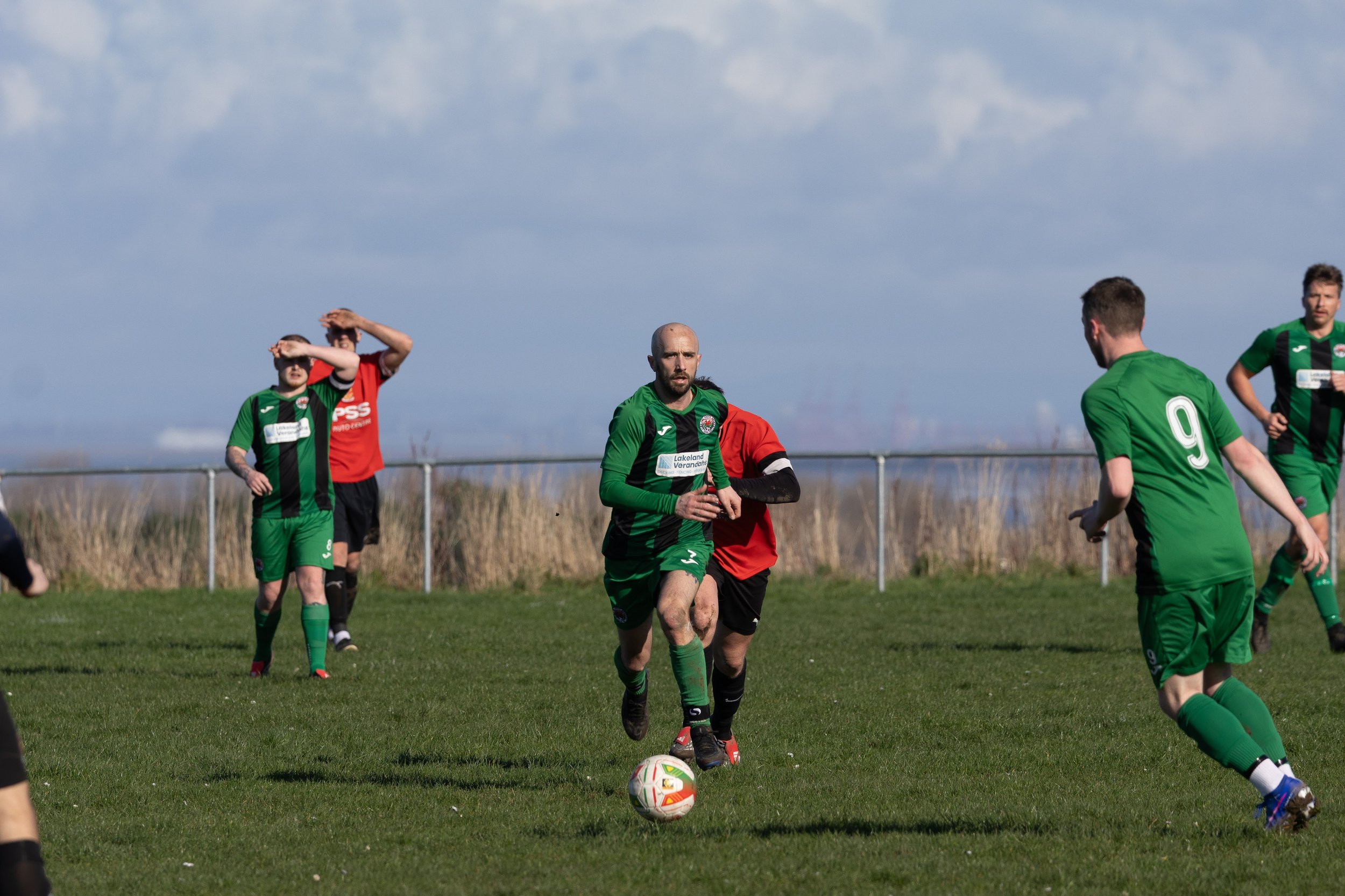 Soccer players on a field, with one in green chasing the ball while others are in background, under cloudy sky.