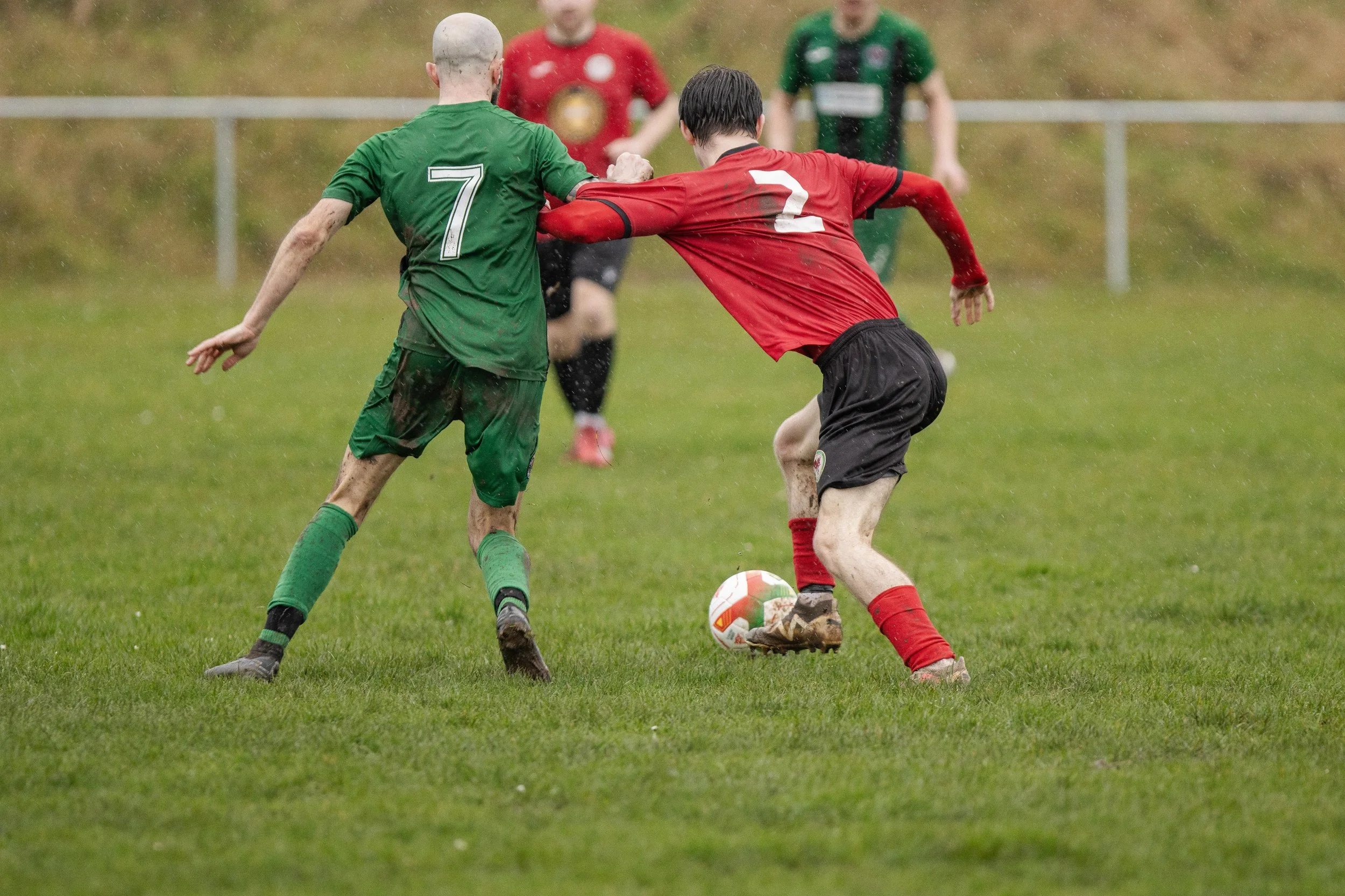 Two soccer players battling for ball on wet grass field, one in green and one in red, with other players in background.