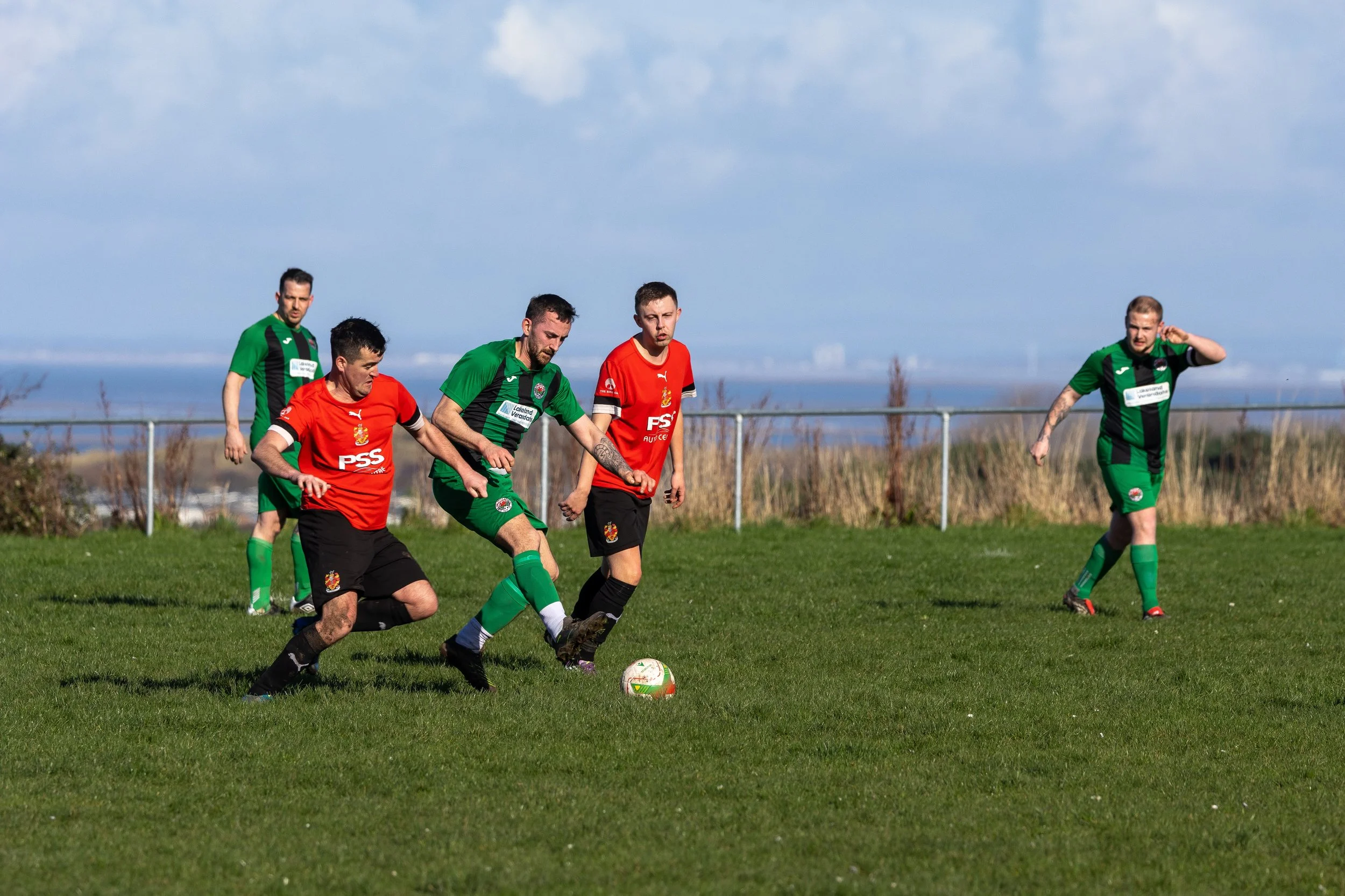 Soccer players in green and red jerseys on a grassy field during a game, with a clear sky and distant landscape in the background.