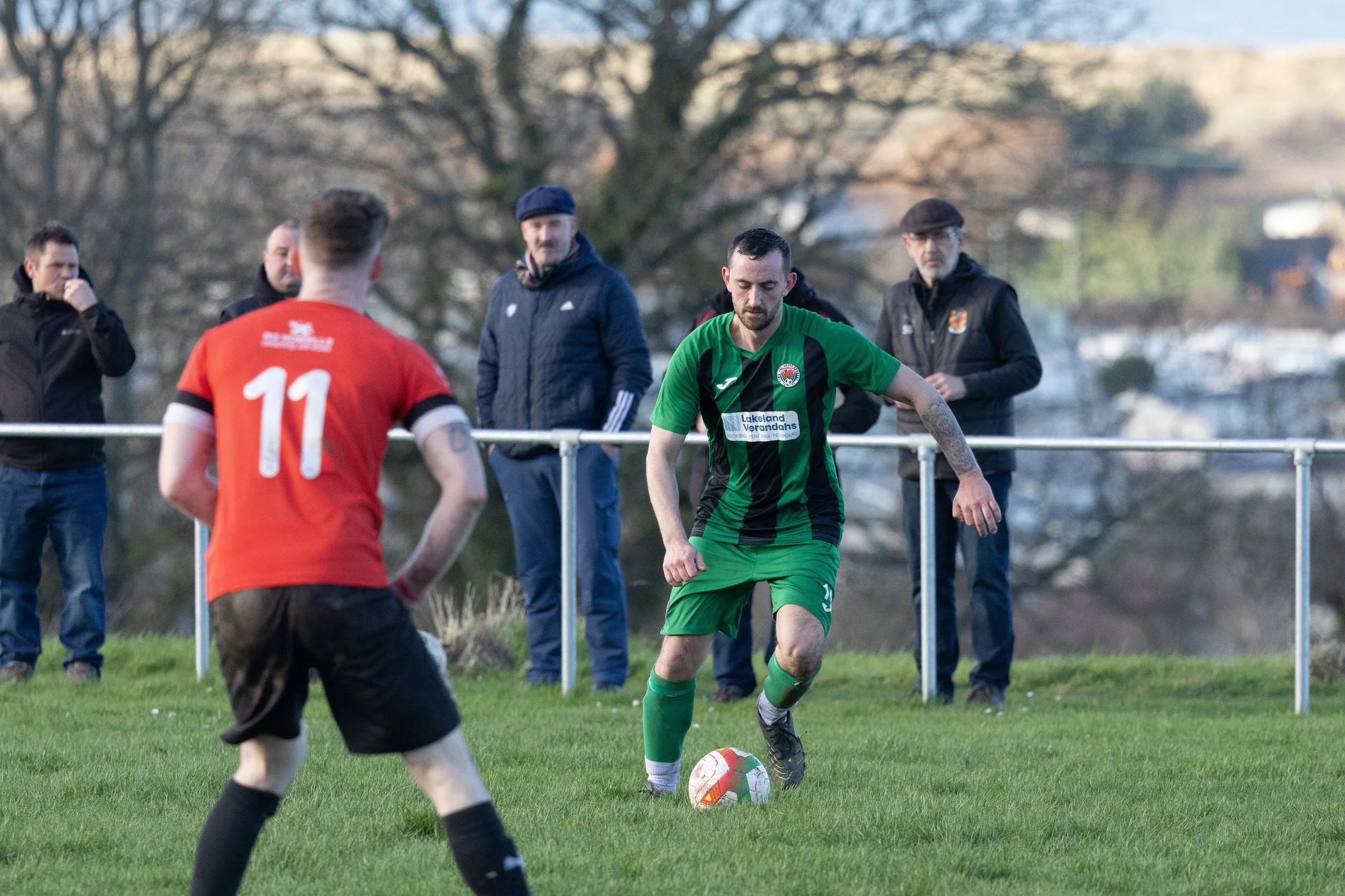 A soccer game in progress on a grassy field with a player in a green and black uniform preparing to kick the ball, while another player in a red and black uniform faces him. Several spectators are watching behind a barrier, with trees and buildings i