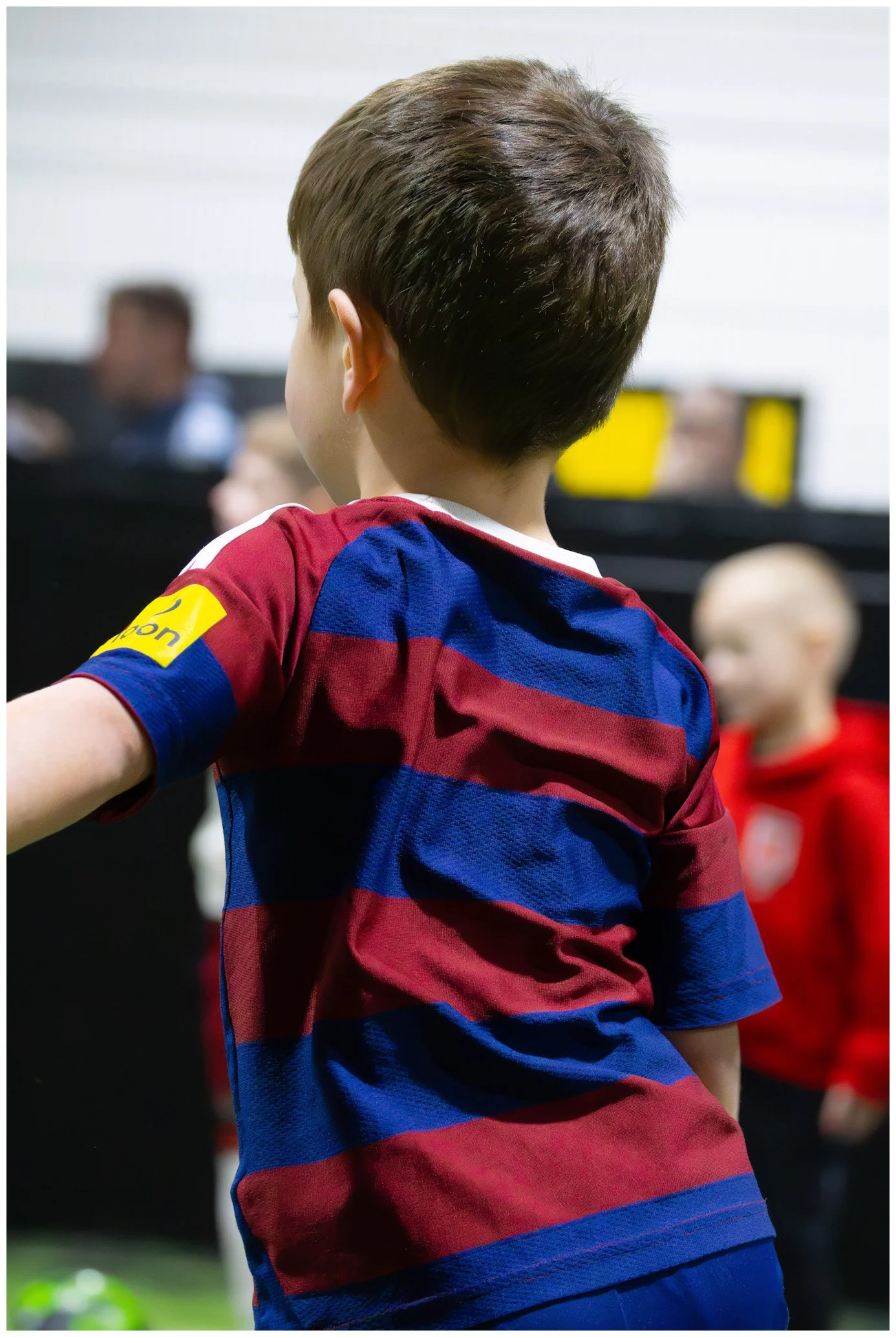 Back view of young boy wearing a red and blue striped soccer jersey with a yellow team patch, standing among other children in an indoor sports setting.