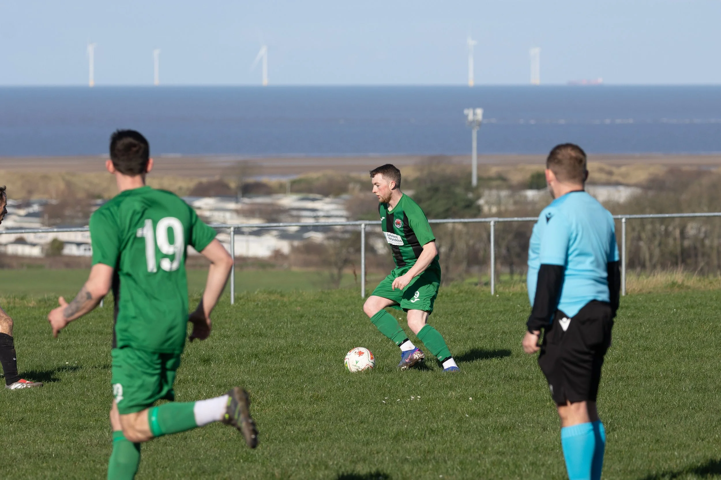 Soccer players in green jerseys on a grassy field near a body of water, with wind turbines in the distance.
