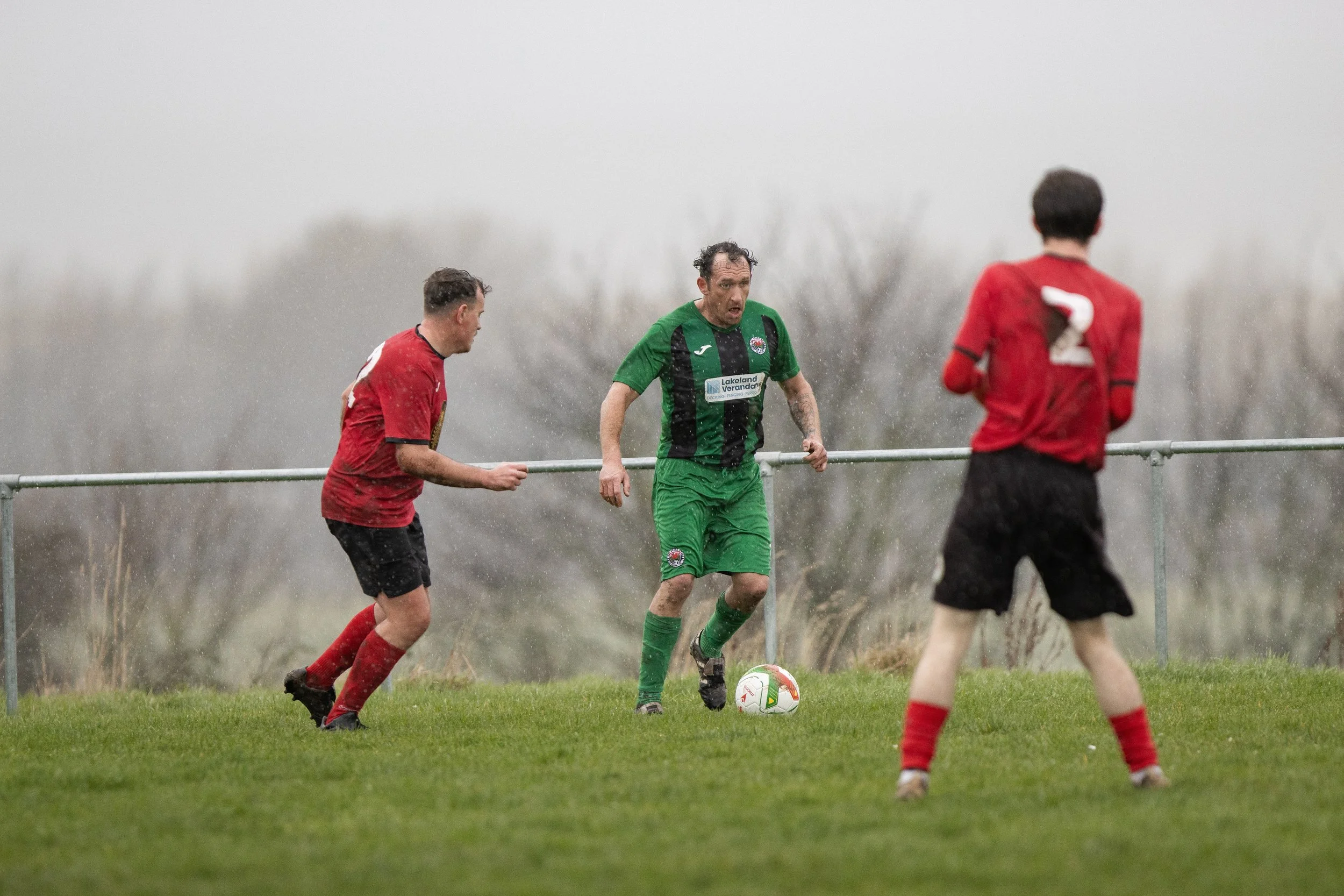 Three soccer players playing on a rainy day, one in a green uniform with a ball at his feet and two in red uniforms approaching him, on a grassy field with blurred trees in the background.