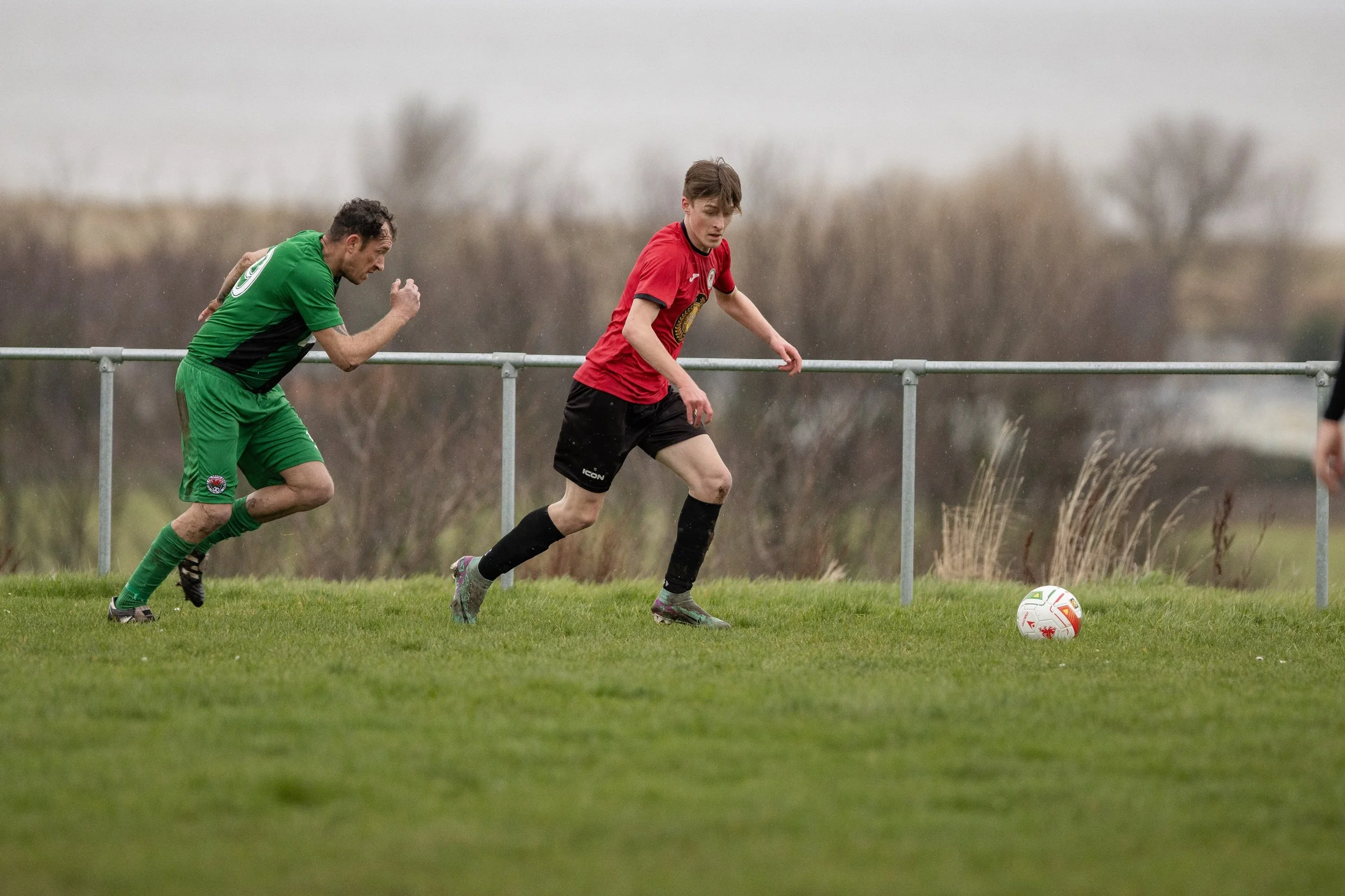 Two soccer players chase a ball on a grassy field with a metal railing and leafless trees in the background, on a cloudy day.