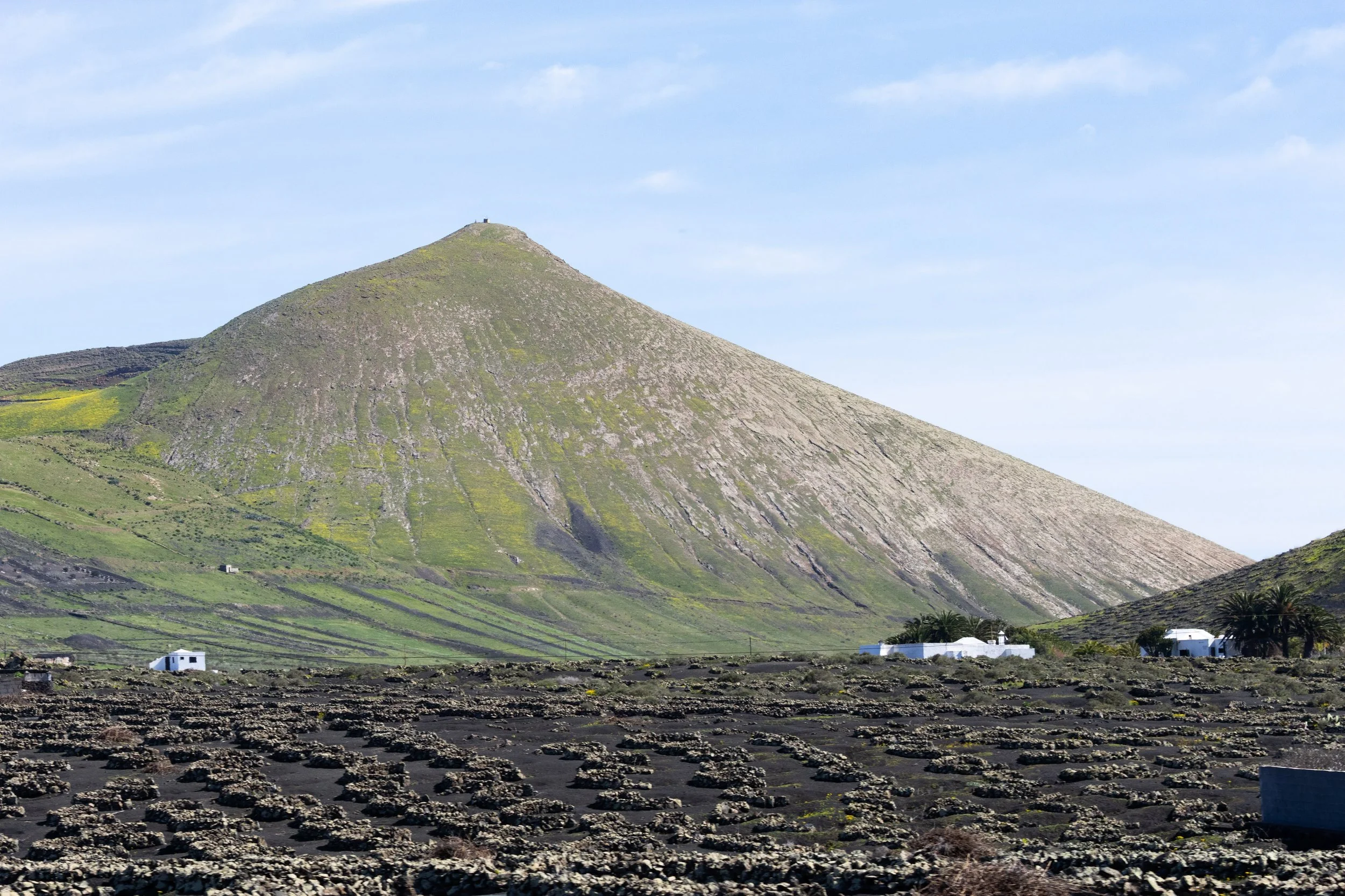 A large, steep, volcanic mountain with patches of green vegetation, with small white buildings and palm trees at the base, under a partly cloudy sky.
