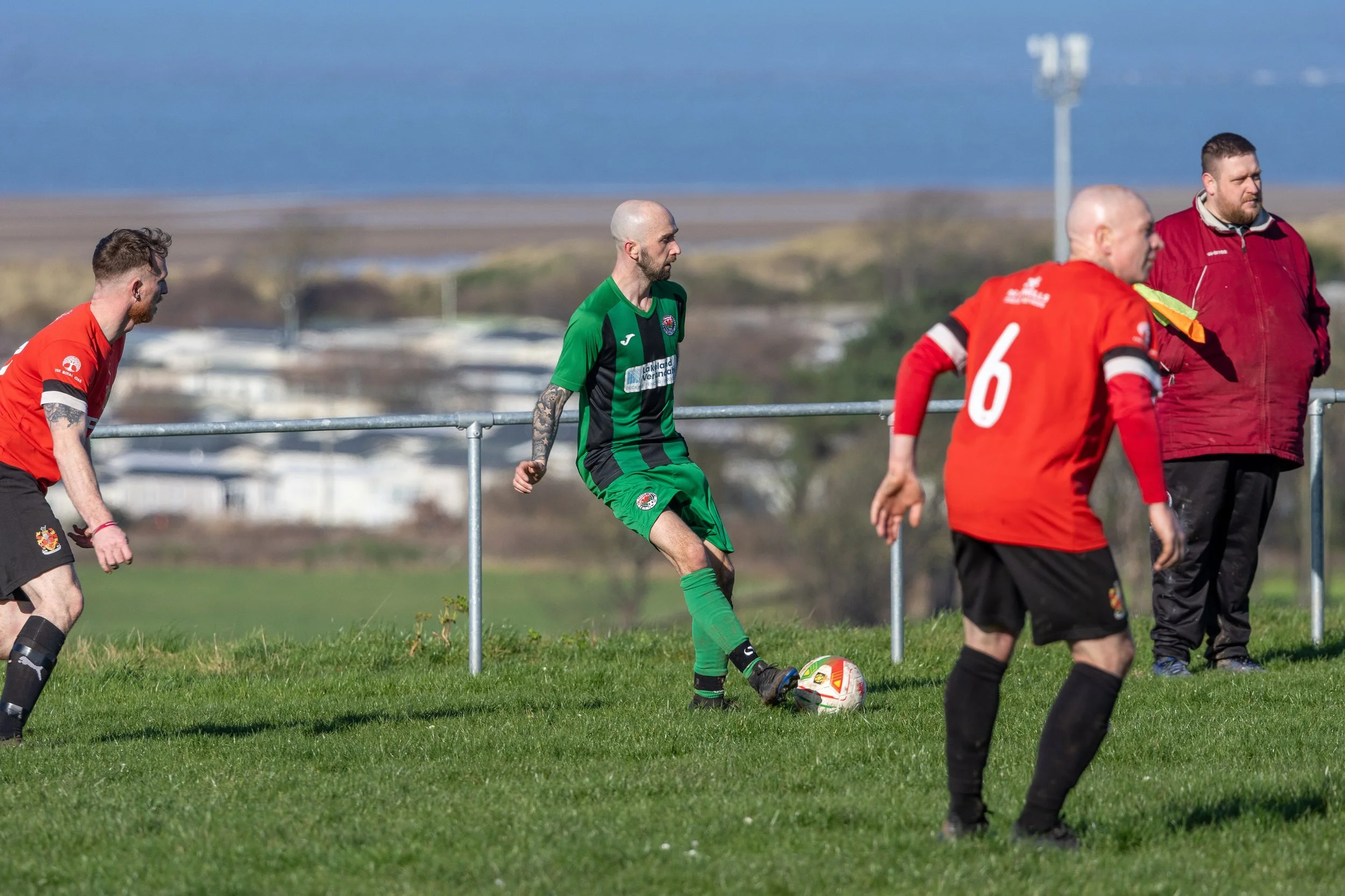 Soccer players on a grassy field, with a man in a green uniform controlling a soccer ball, and others in red uniforms nearby. There is a landscape with water and buildings in the background.