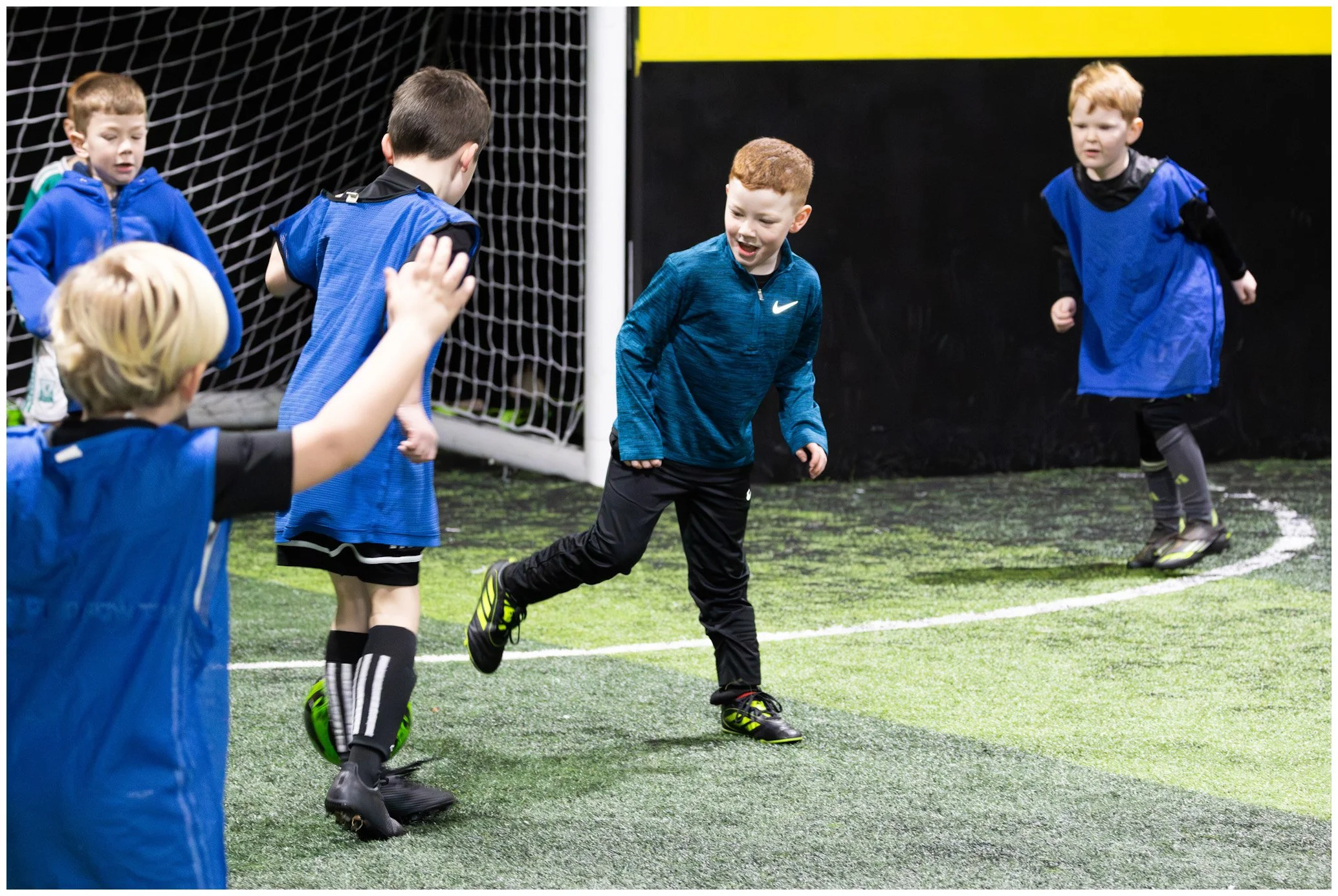 Young boys playing soccer indoors on a turf field, some in blue pinnies, with a goalpost and black wall in the background.