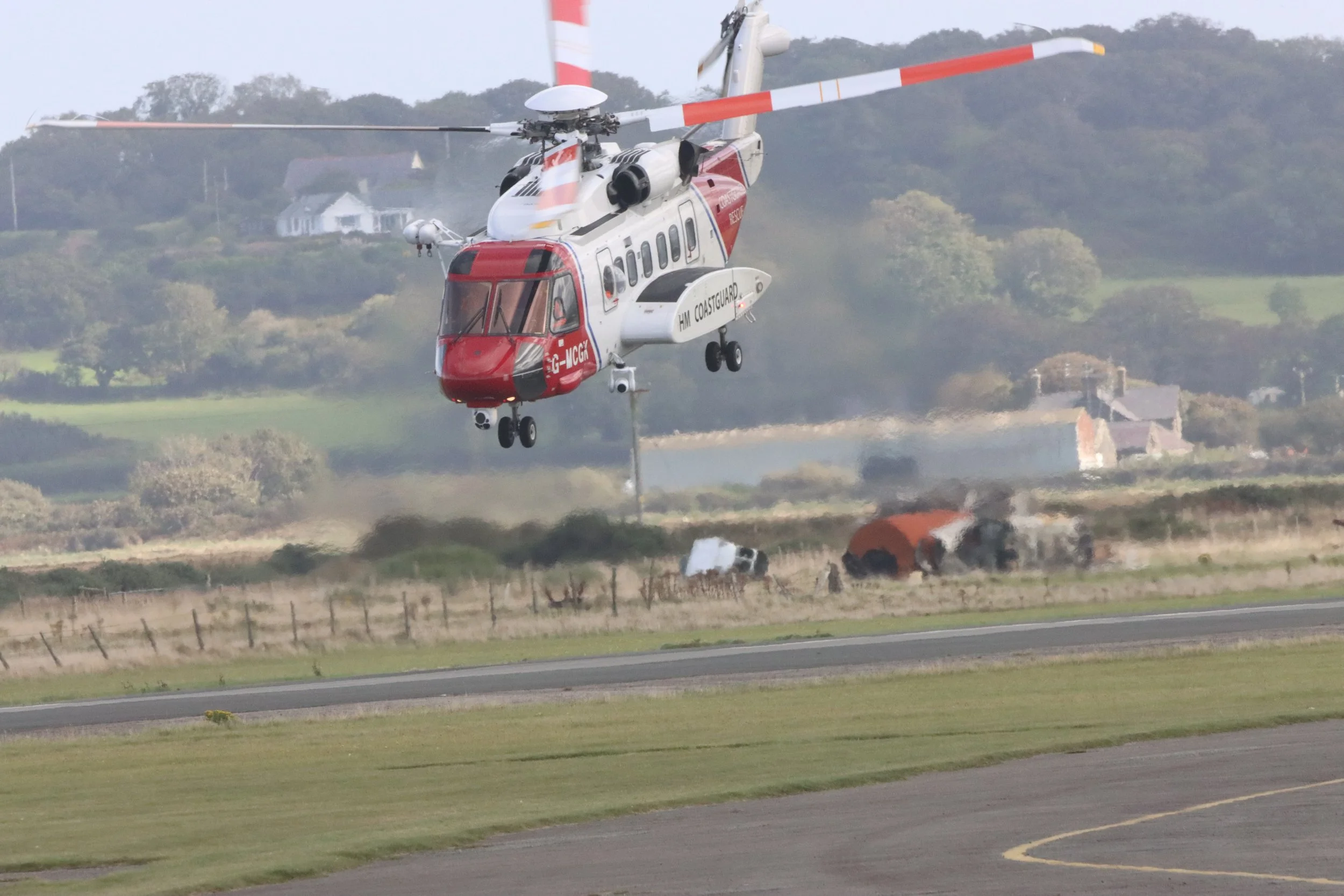 A rescue helicopter in flight after collision with a vehicle; smoke rises from the wreckage burned on the ground beneath it, with a rural landscape and houses in the background.
