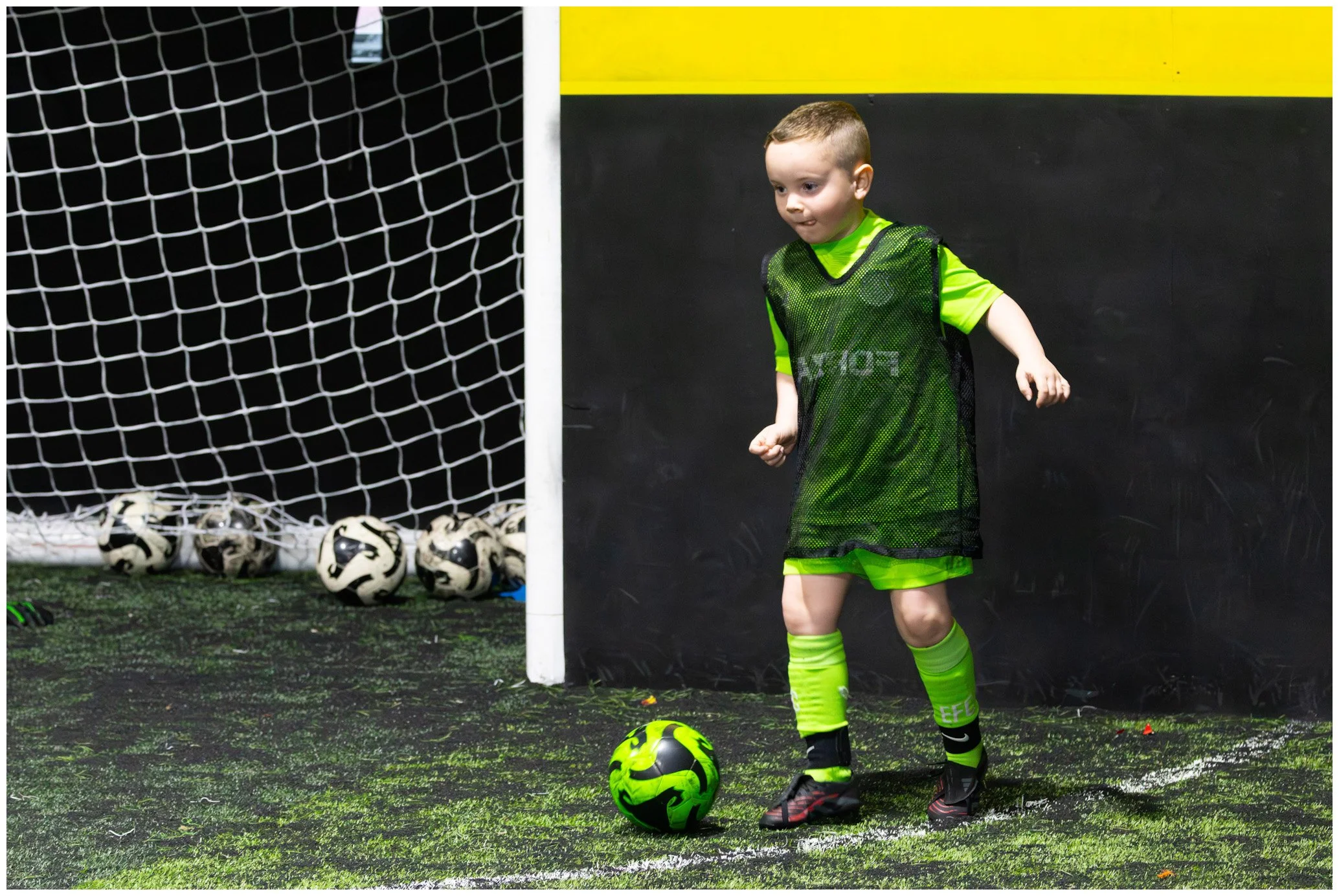 Young boy in neon green sports gear standing next to a soccer ball on a turf field, with a soccer goal and several soccer balls in the background.