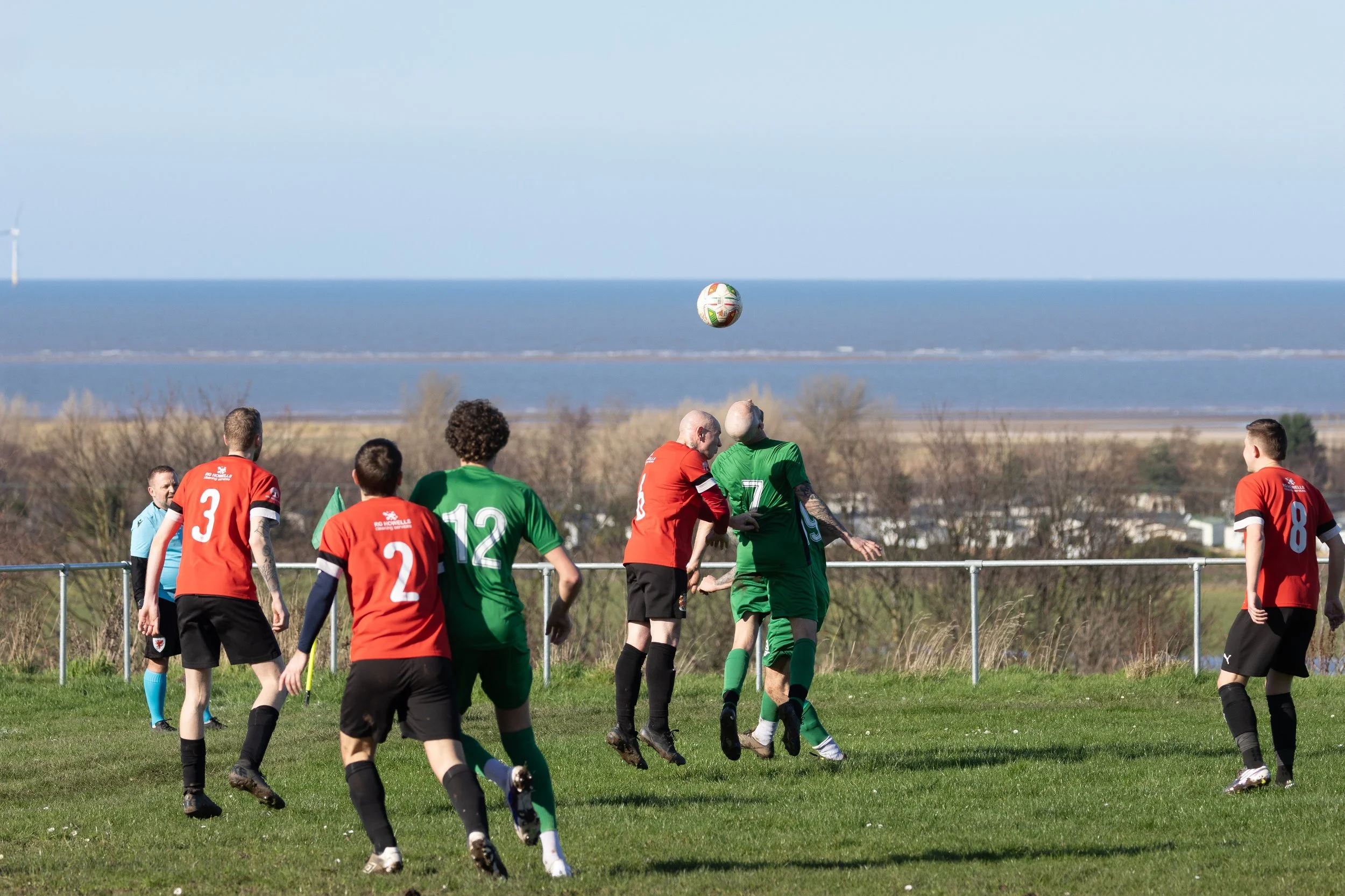 Soccer game with players in red and green jerseys jumping and trying to head the ball, with a seaside view in the background.
