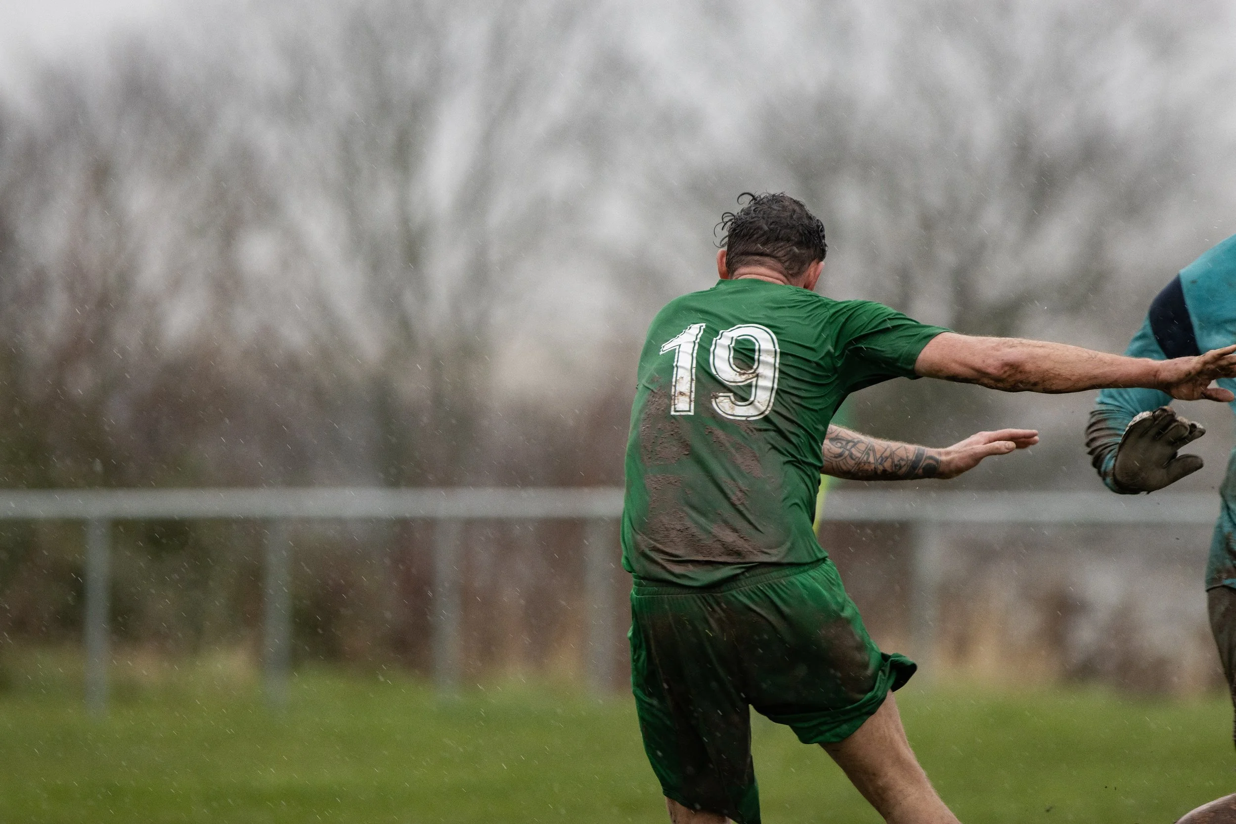 A soccer player wearing a green jersey with the number 19 is kicking a ball in the rain during a match.
