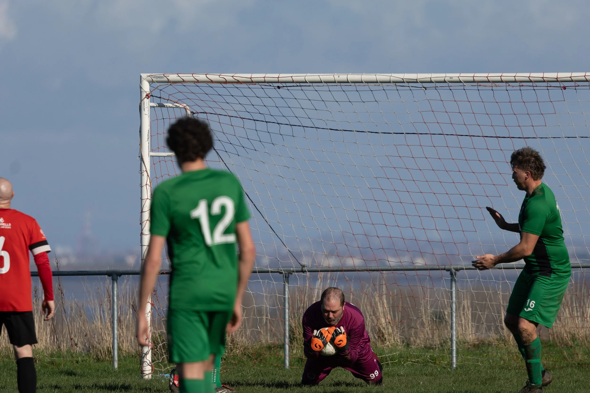 A soccer goalkeeper in a purple uniform catching or handling the ball during a match, with players in green and red uniforms nearby on a grassy field.