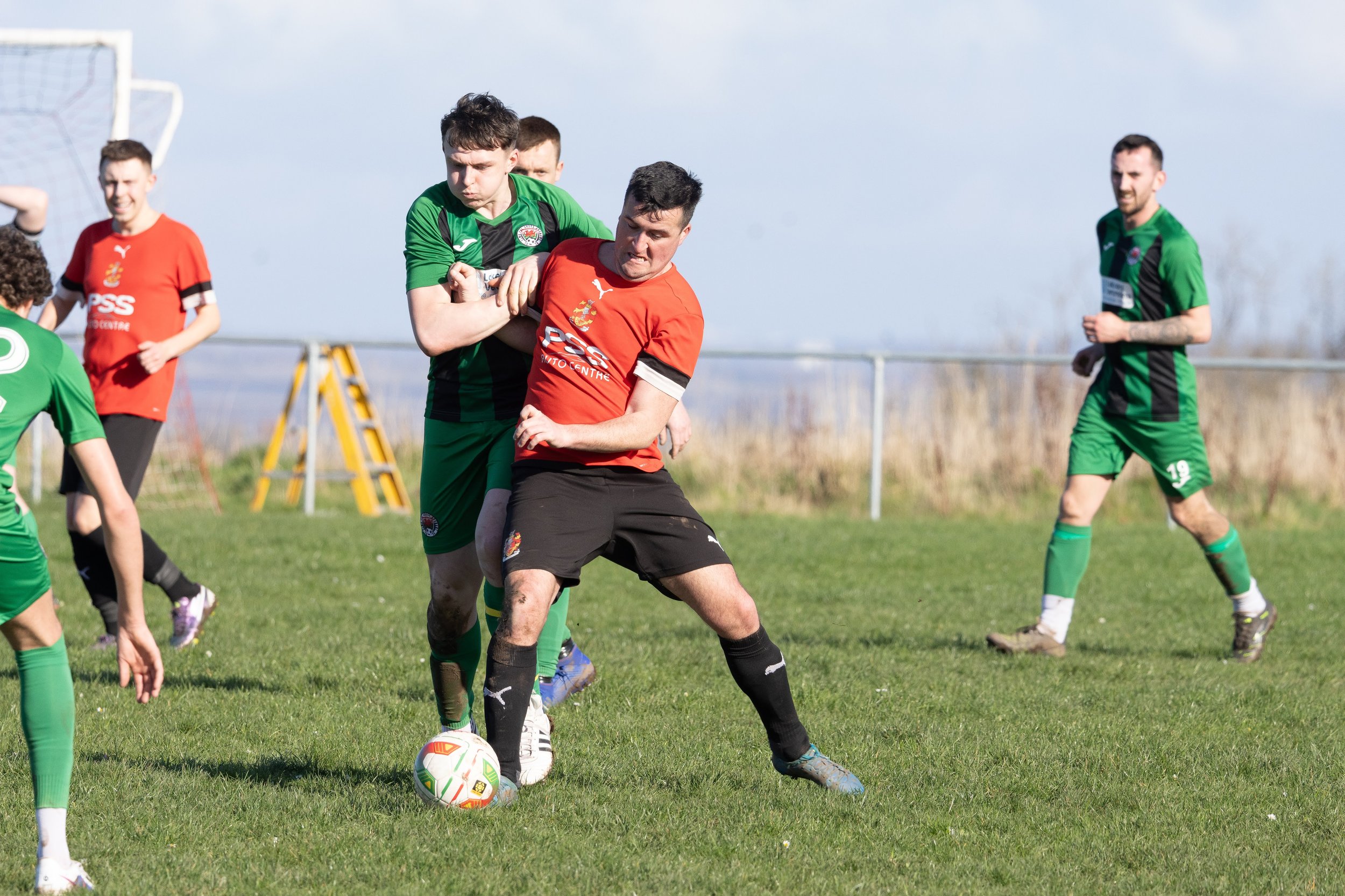 Soccer players in green and red uniforms competing for the ball on a grassy field under a partly cloudy sky.