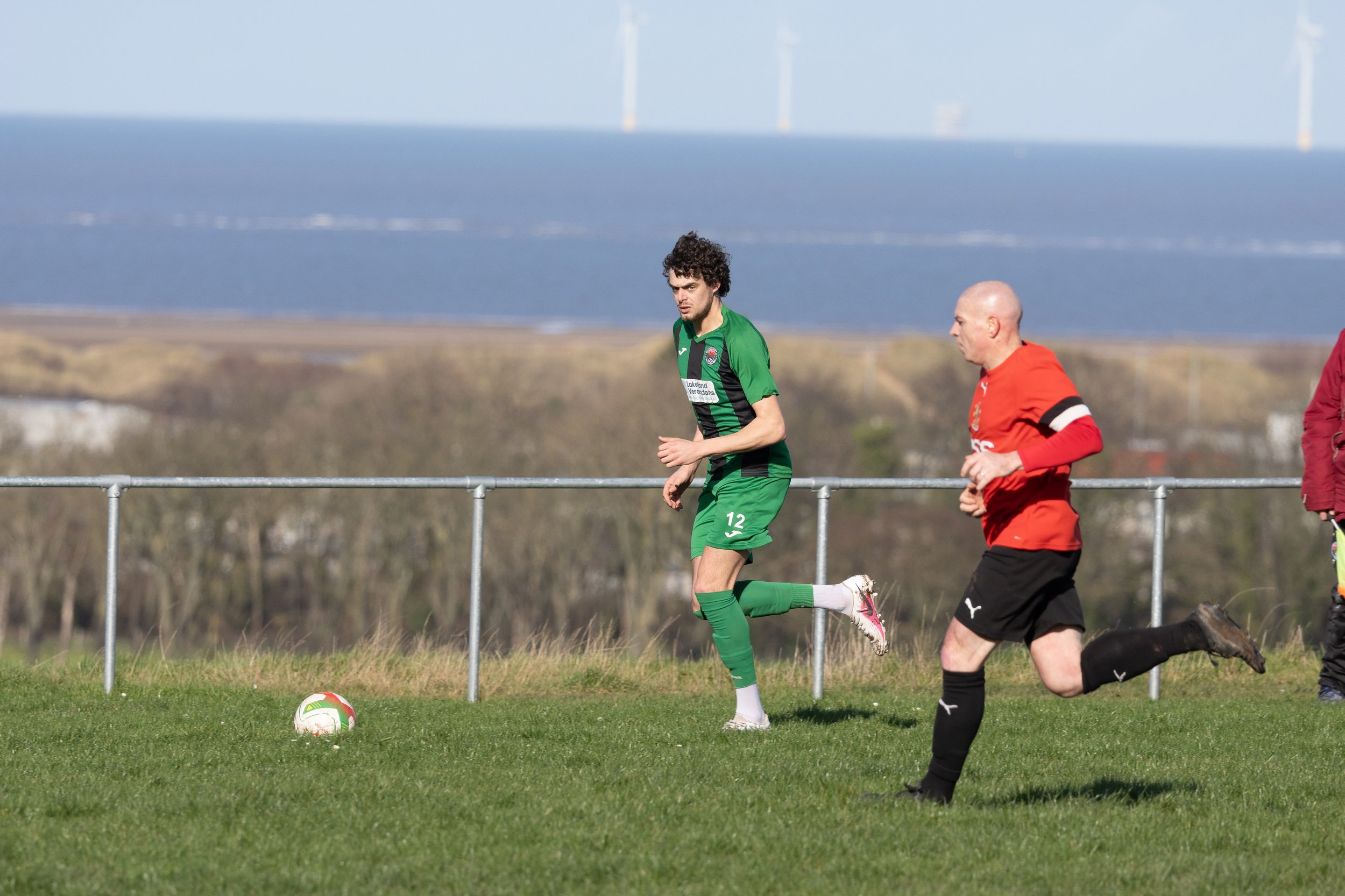 Two soccer players on a grassy field during a game, with a soccer ball nearby and a body of water with sailboats in the background.