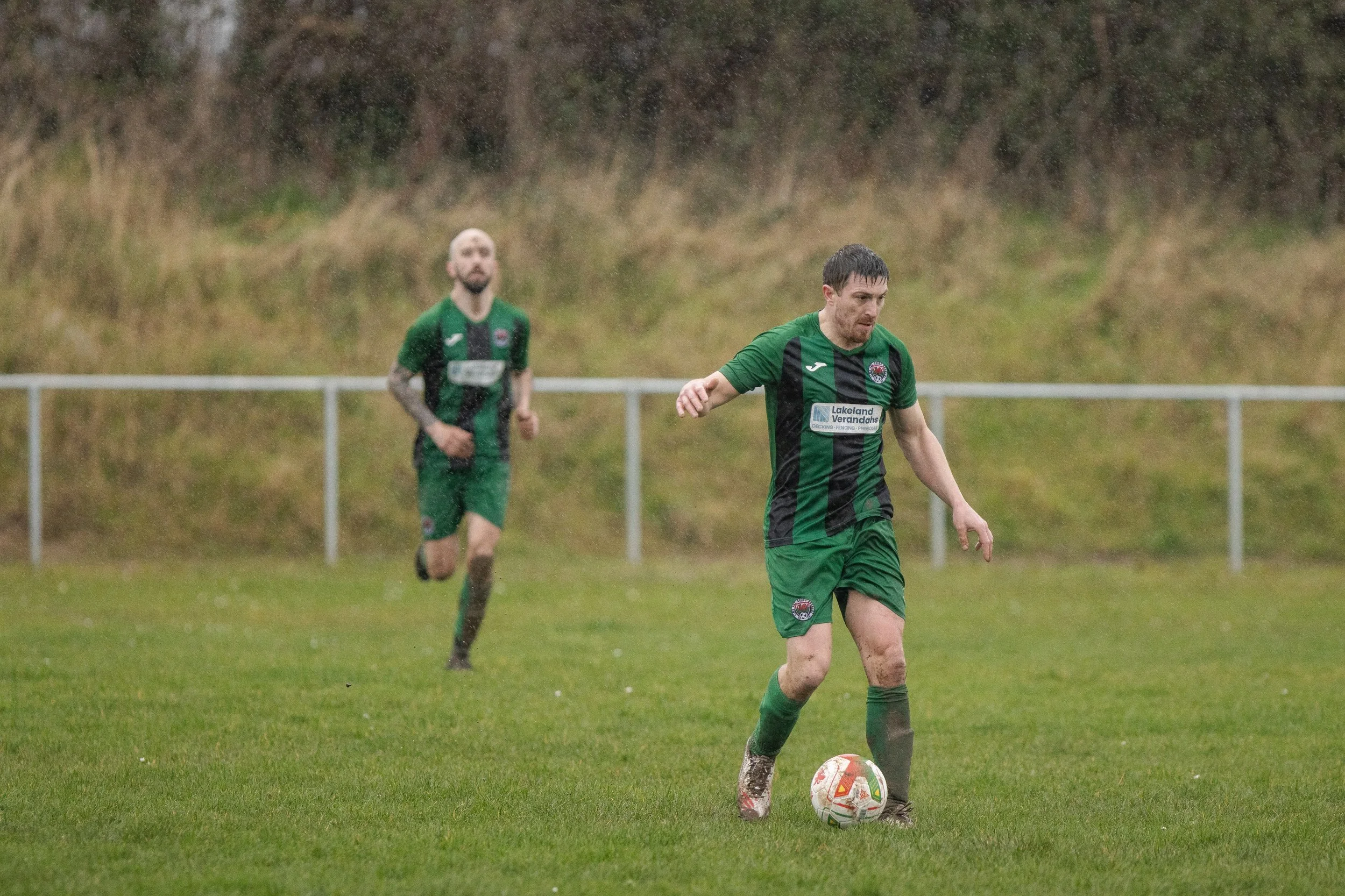 Two soccer players in green and black uniforms playing on a grassy field in rainy weather, with one player dribbling the ball and the other running in the background.