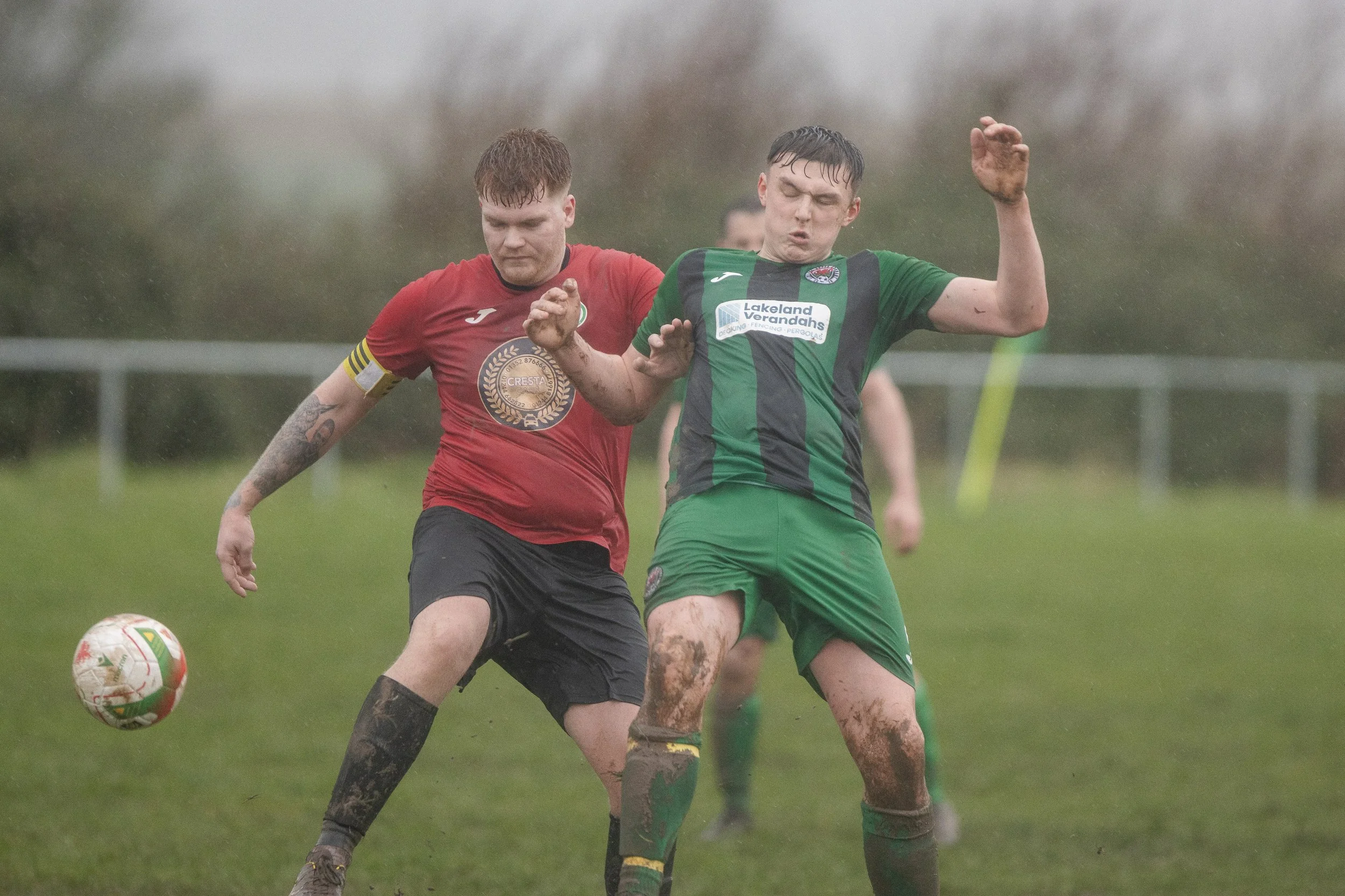 Two soccer players compete for the ball on a muddy field during a rainy game, one wearing a red jersey and the other wearing a green and black jersey.