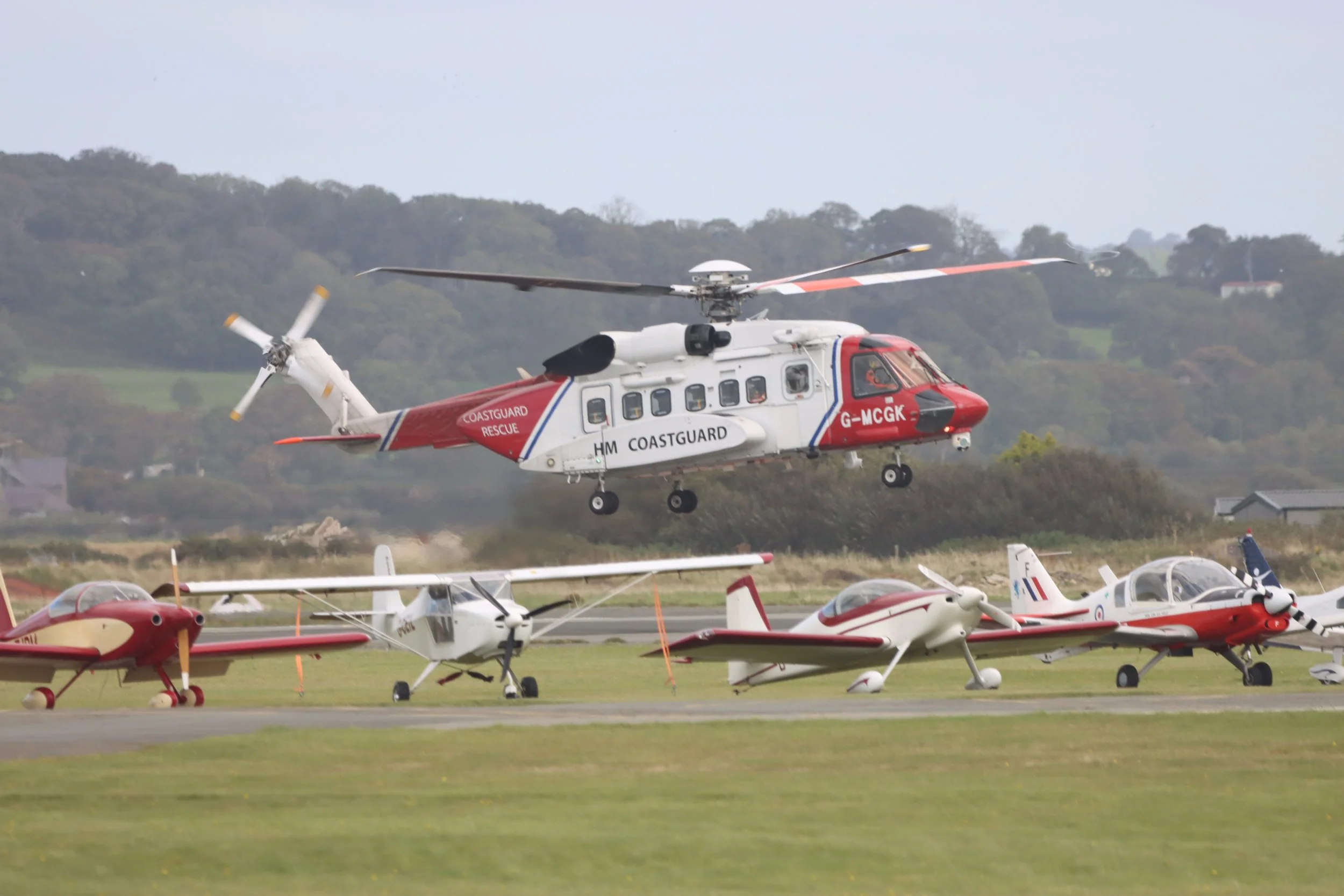 A helicopter with the text 'HM Coastguard' and 'Rescue' on the side, flying above small airplanes parked on a runway, with a hilly landscape in the background.