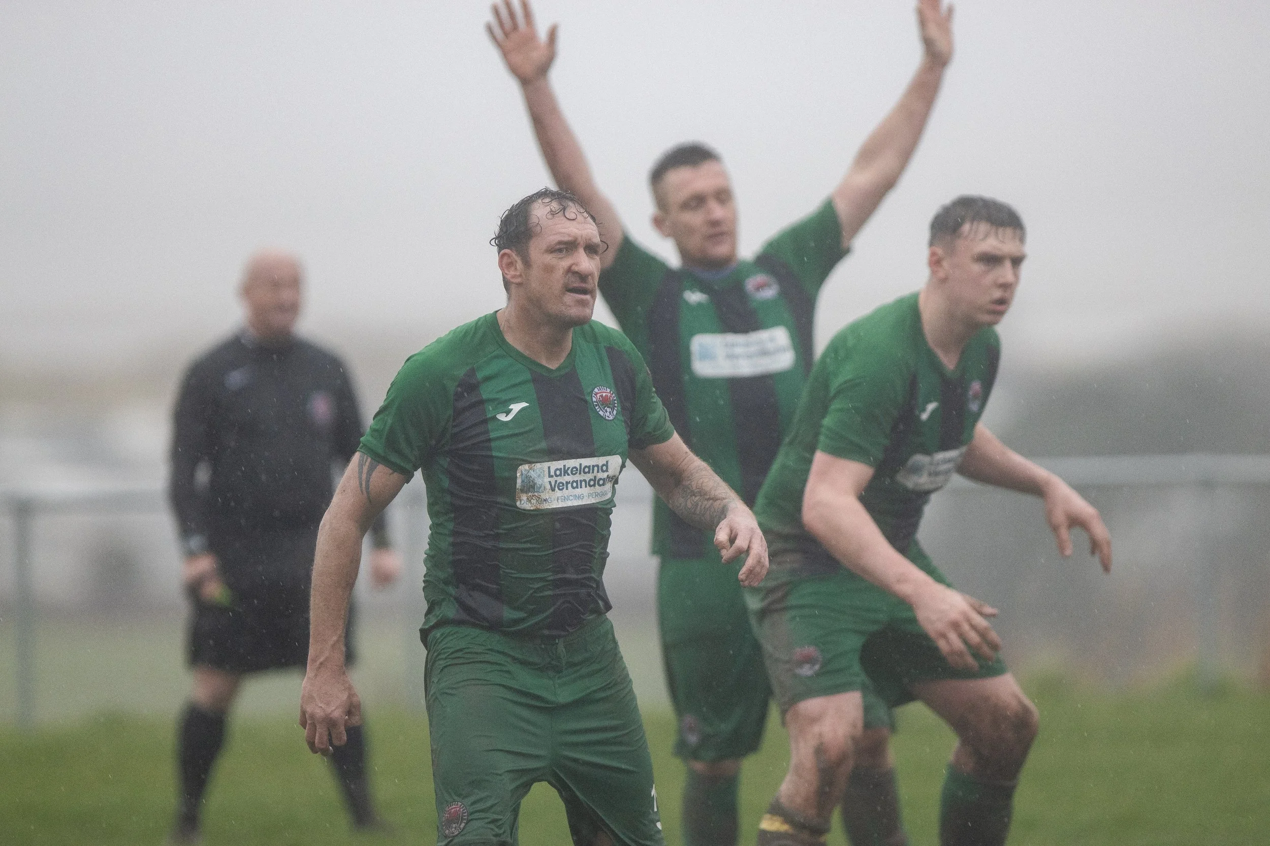 Football players in green jerseys and mud, reacting during a game on a foggy field, with a referee in black in the background.