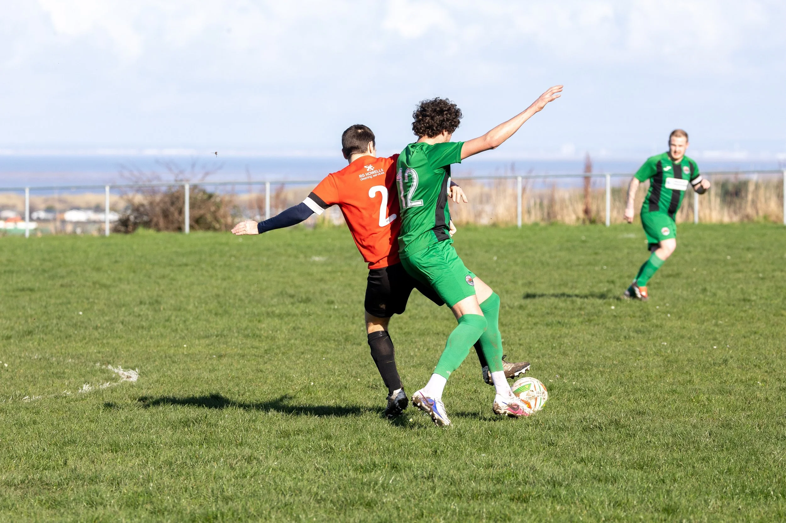Two soccer players competing for the ball on a grassy field, with a third player running towards them in the background. The player in green is shielding the ball while the player in red tries to challenge.