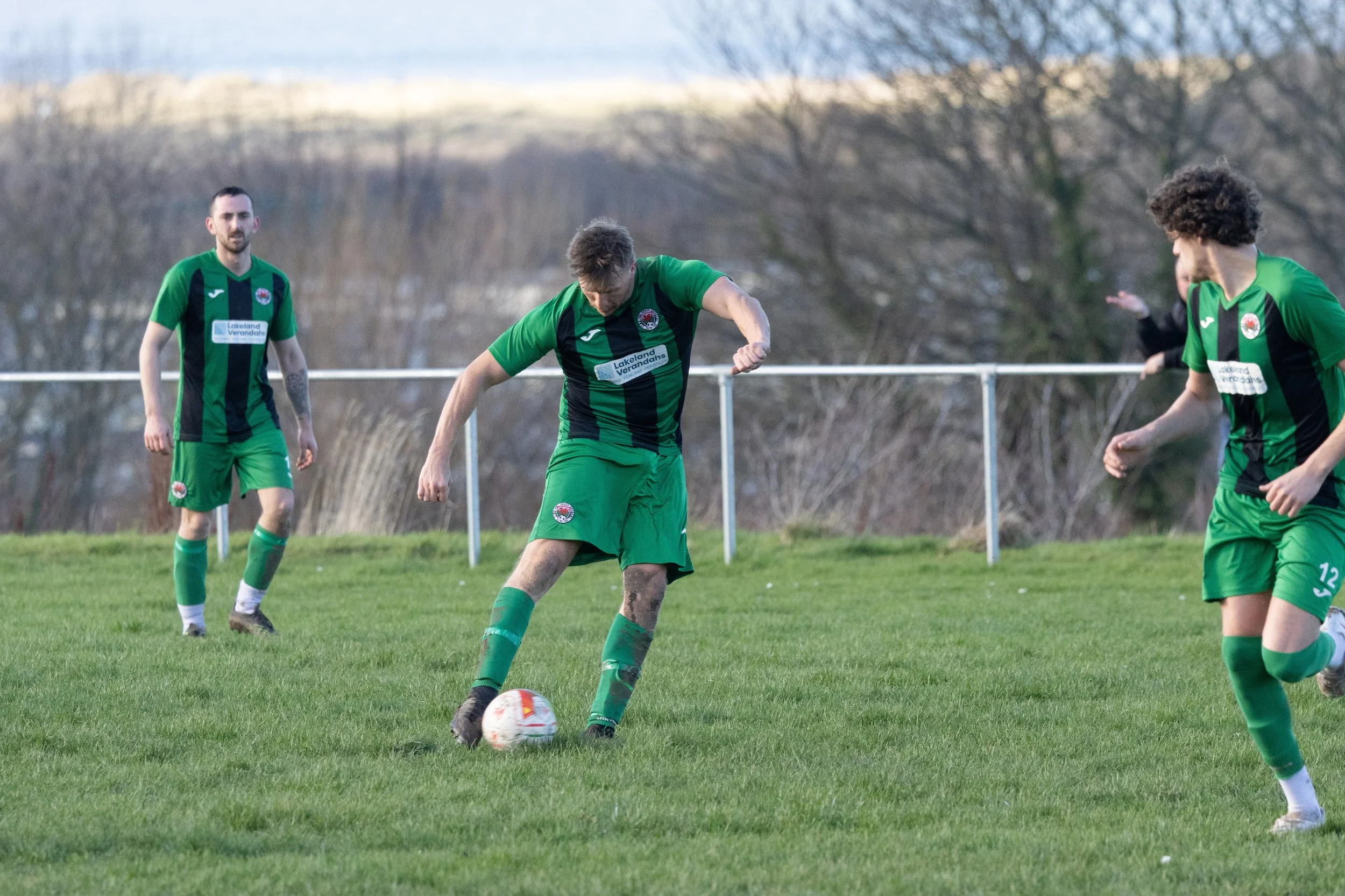 Soccer players in green and black uniforms on a grassy field during a match, with one player preparing to kick the ball while others watch.
