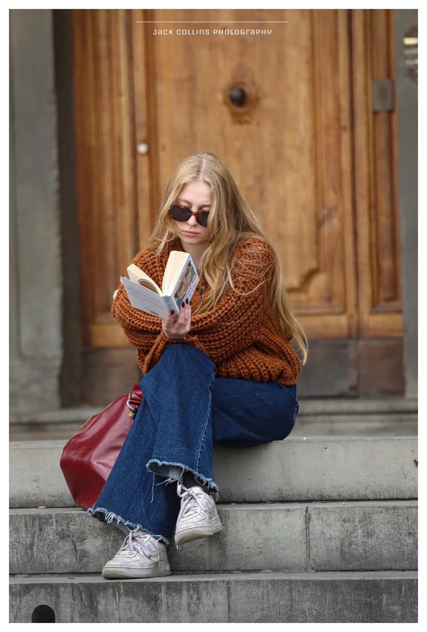 A young woman with long blonde hair wearing sunglasses, a chunky brown sweater, and frayed jeans, sitting on stone steps and reading a book, with a large red bag beside her, in front of a wooden door.