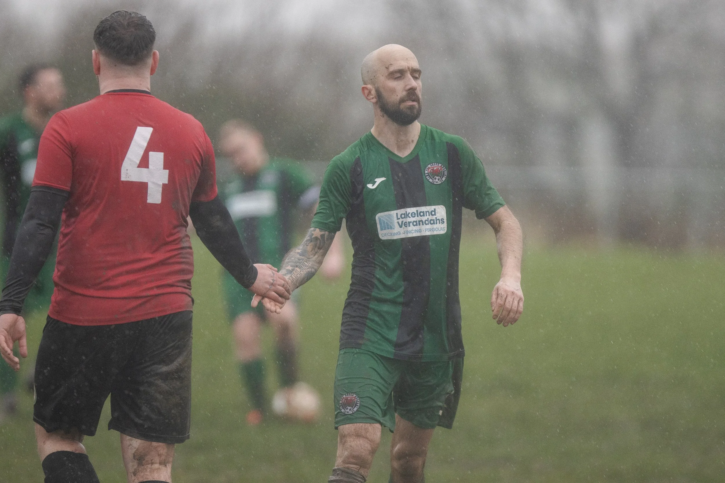 Two soccer players holding hands during a game in rainy weather. One player wears a red and black uniform with the number 4, and the other wears a green and black uniform with a tattooed arm.