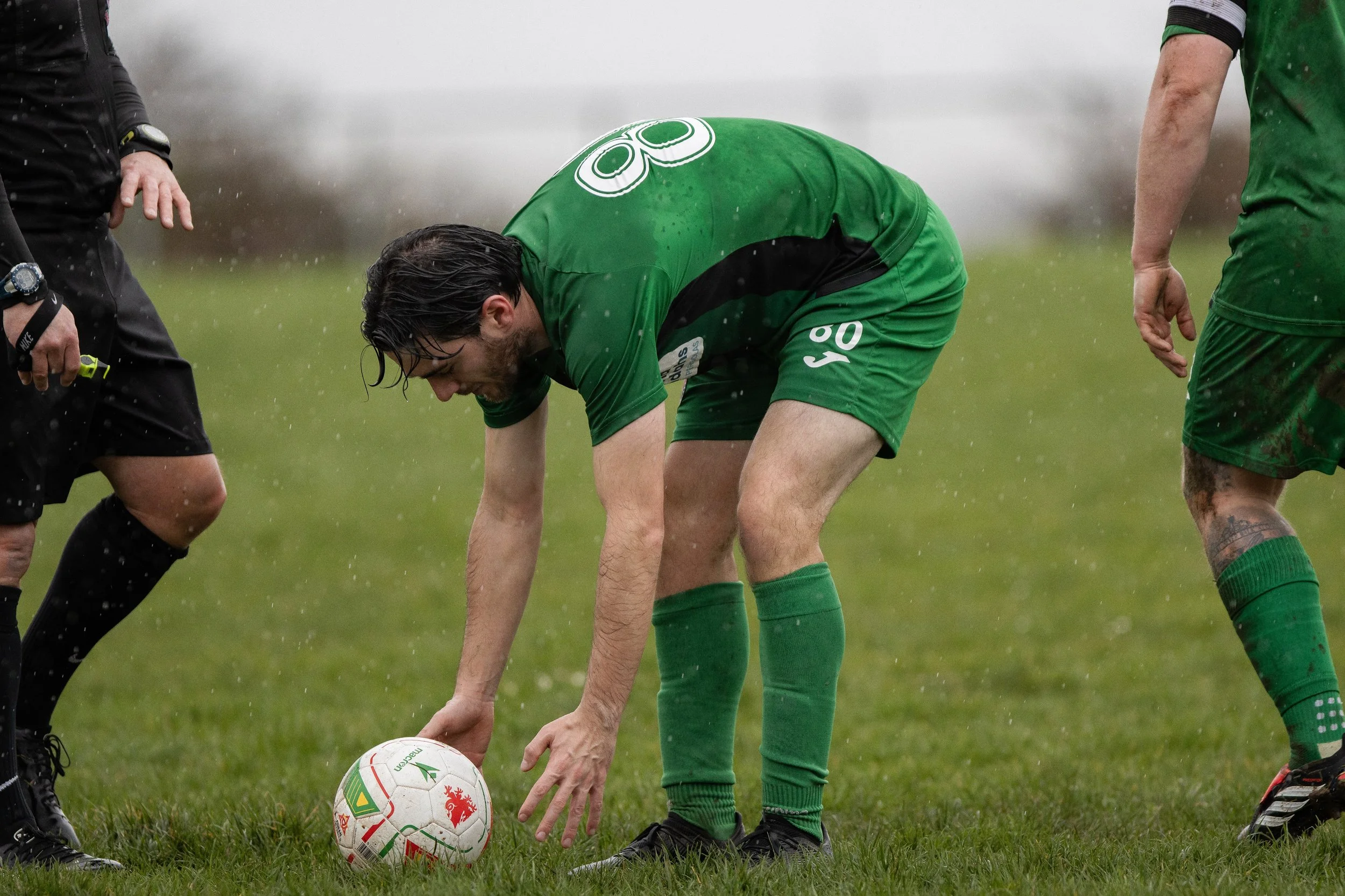 Soccer player in green uniform bent over to place a ball on the field, with rain falling and other players nearby.