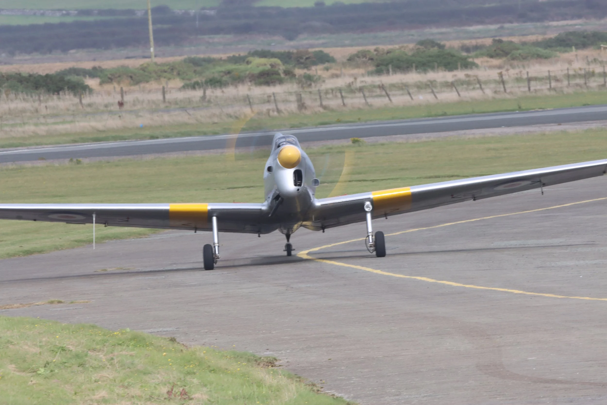 A silver vintage fighter aircraft taxis on a runway with a yellow nose cone and yellow markings on its wings, surrounded by grassy fields and distant hills.