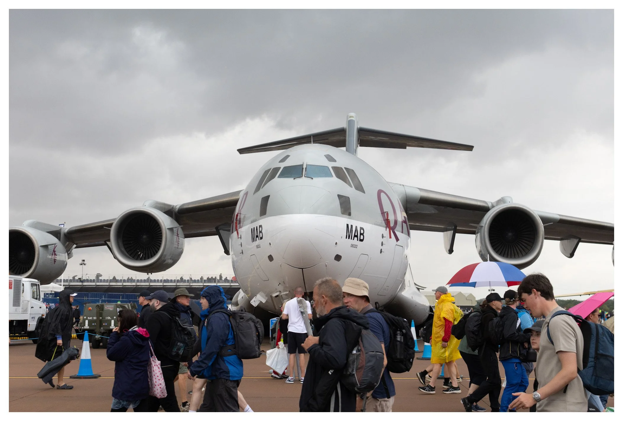 People walking near a large military cargo aircraft on an airfield with cloudy skies.